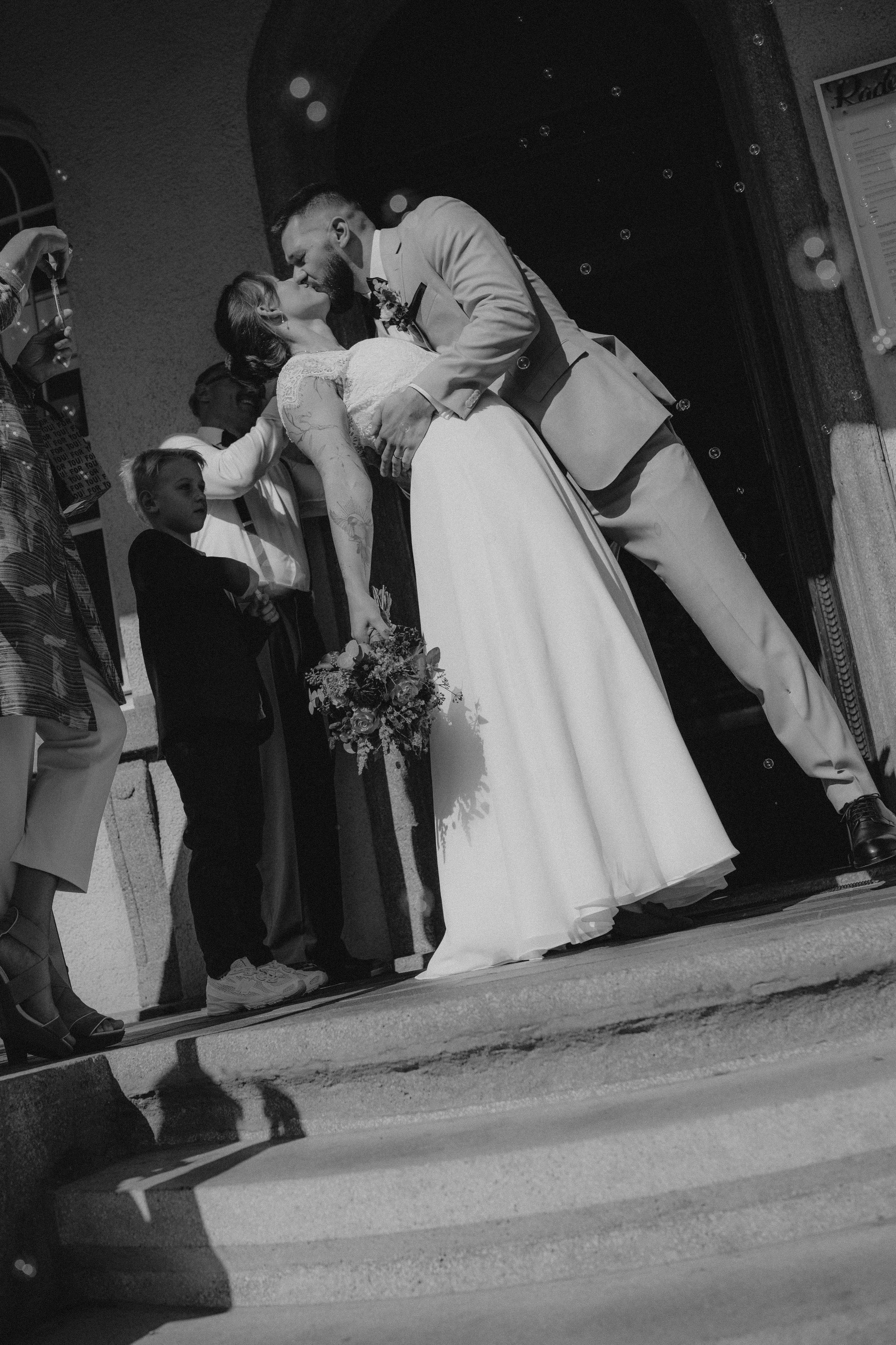 Ein Paar bei einer Hochzeitszeremonie in einer Kirche, der Bräutigam küsst die Braut, umgeben von Gästen.