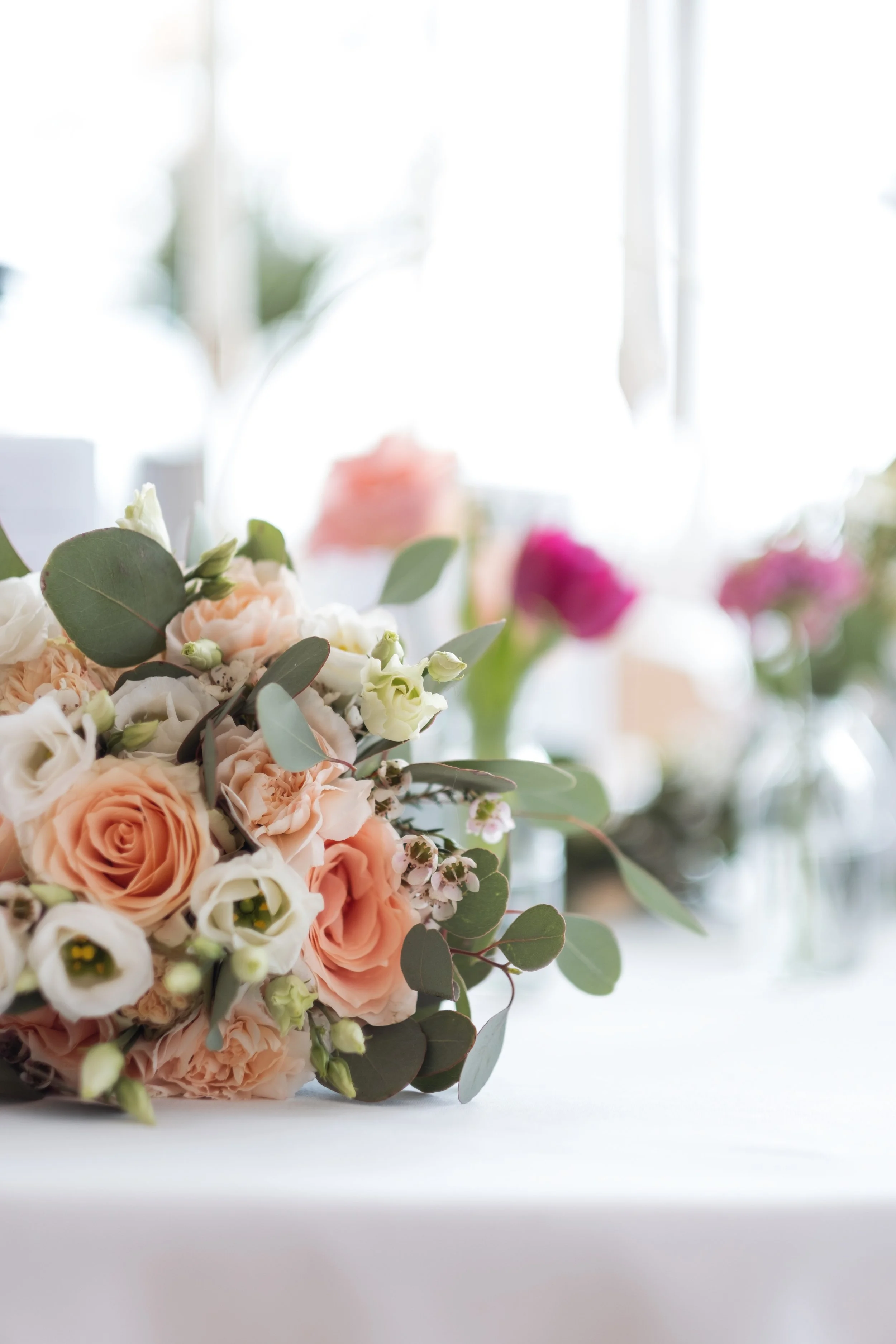Blumenstrauß mit peachfarbenen Rosen und Eukalyptus auf einem weißen Tisch, im Hintergrund verschwommene weitere Blumen. Natürliche Beleuchtung.