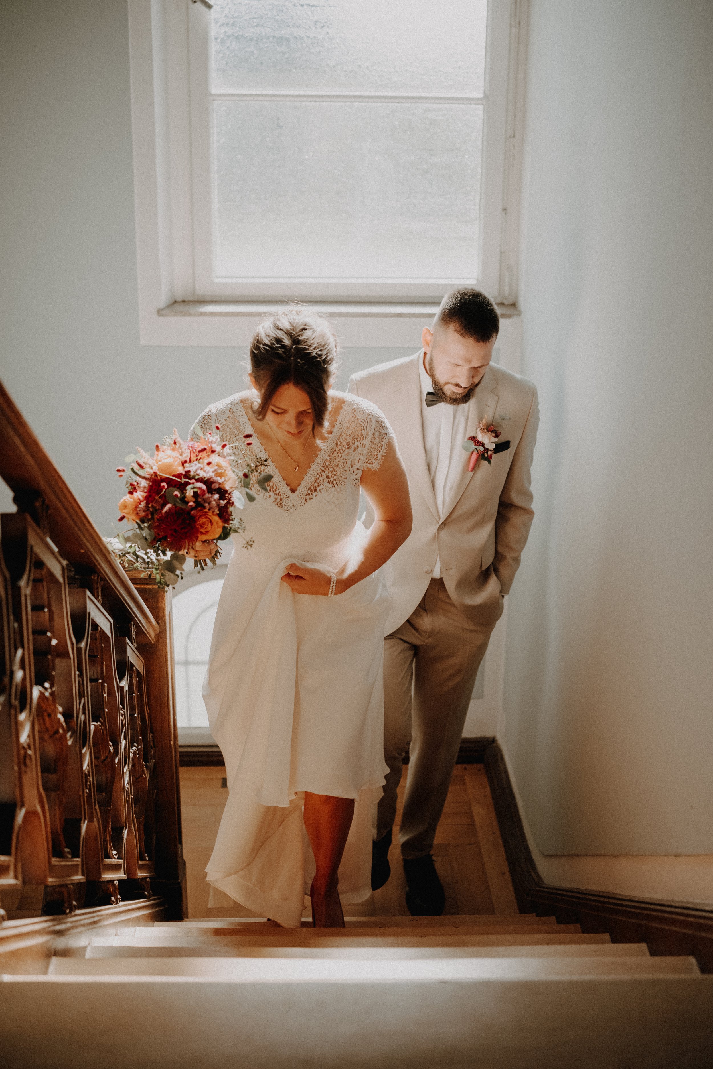 Ein Brautpaar, das die Treppe in einem hell erleuchteten Raum hinaufgeht. Die Braut trägt ein weißes Kleid mit Spitzendetails und hält einen Blumenstrauß, während der Bräutigam einen beige Anzug und Krawatte trägt.