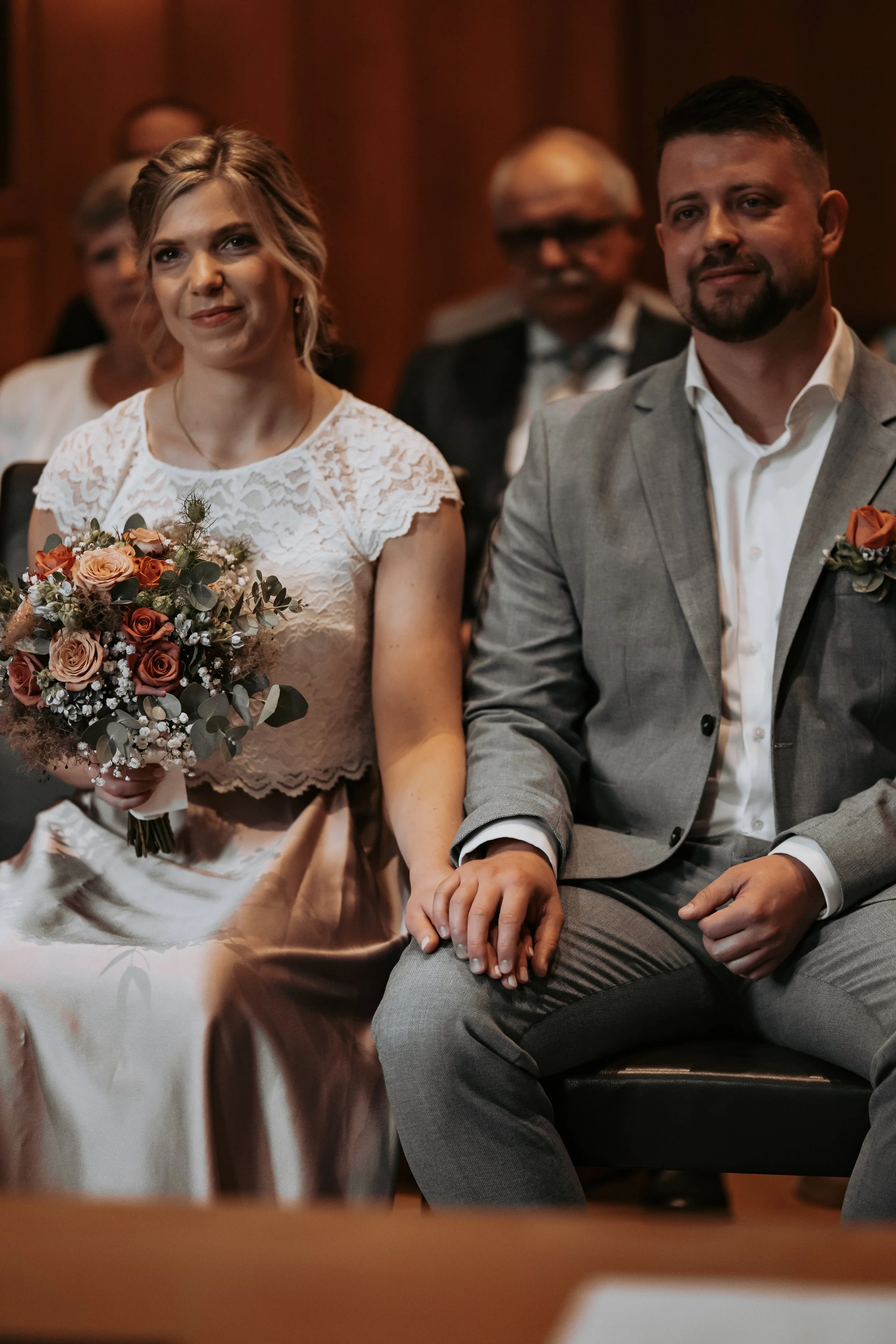 Ein Brautpaar bei einer Hochzeit, die Braut hält einen Blumenstrauß, sie trägt ein weißes Spitzenkleid, der Bräutigam trägt einen grauen Anzug, sie sitzen im Saal, im Hintergrund sind weitere Gäste zu sehen.