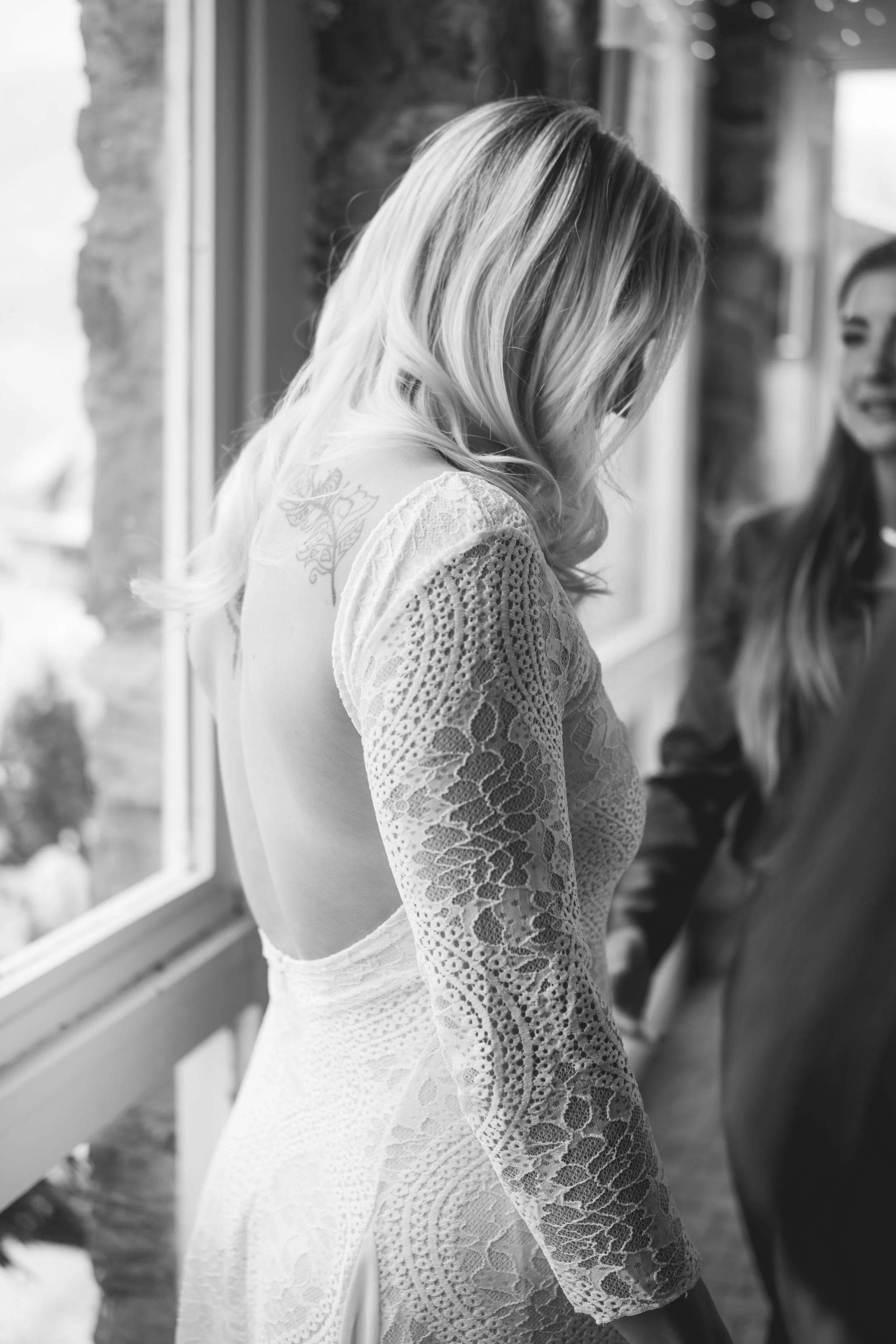 Frau in einem Hochzeitskleid steht neben einer anderen Frau vor einem Fenster, beide lachen.