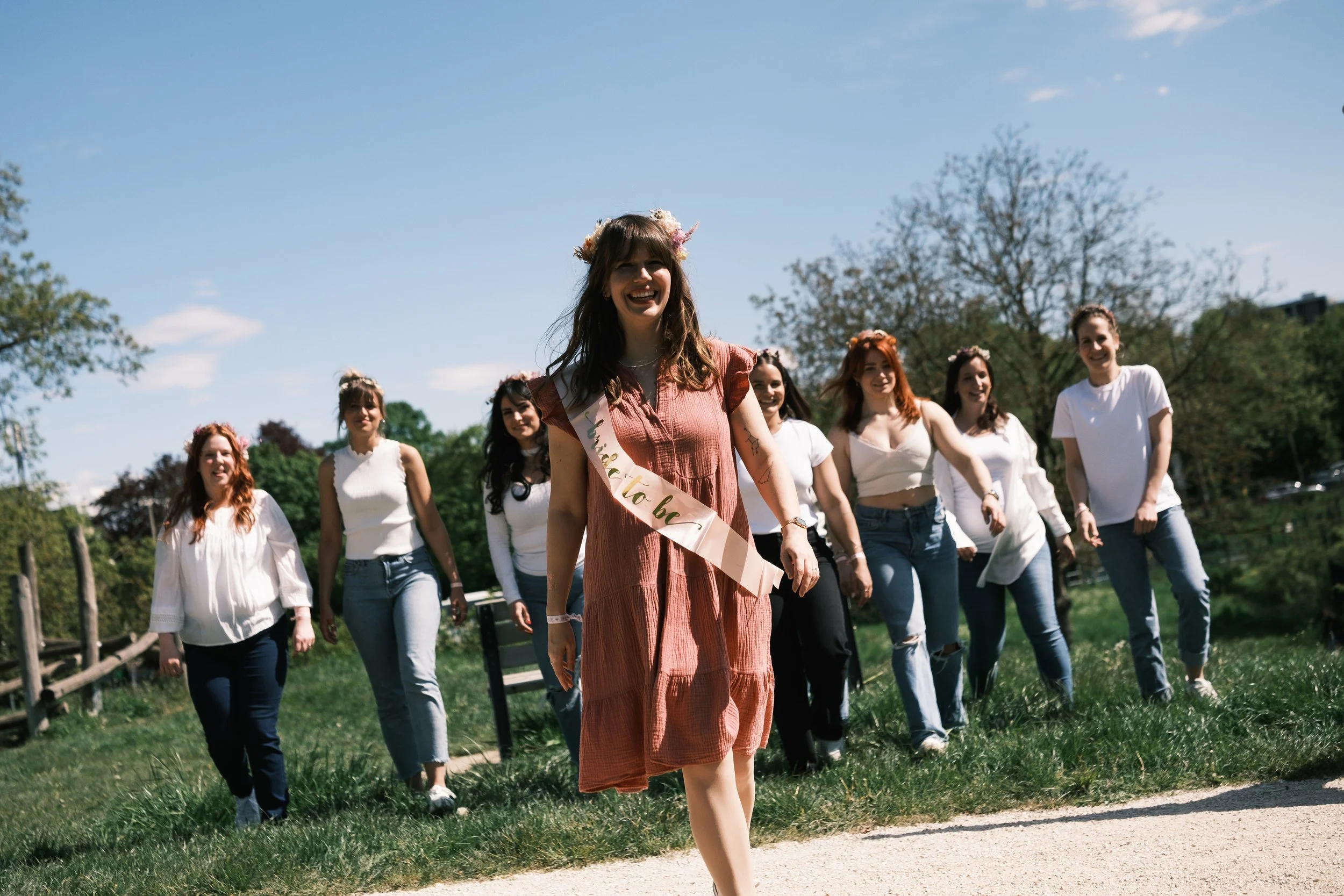 Eine Gruppe Frauen, inklusive der Frau mit einer Banderole, die wahrscheinlich die Braut ist, laufen auf einem Rasen im Freien bei sonnigem Wetter, umgeben von Bäumen.