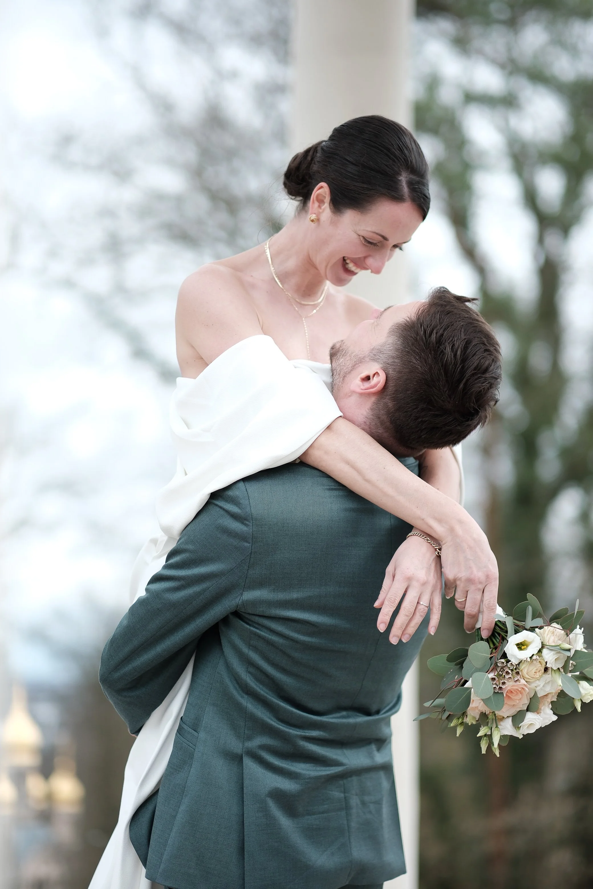 Ein Mann hebt eine Frau hoch, beide lachen und schauen sich verliebt an, bei einer Hochzeit im Freien mit Bäumen im Hintergrund, sie trägt ein weißes Kleid, er trägt einen dunklen Anzug, sie hält einen Blumenstrauß.