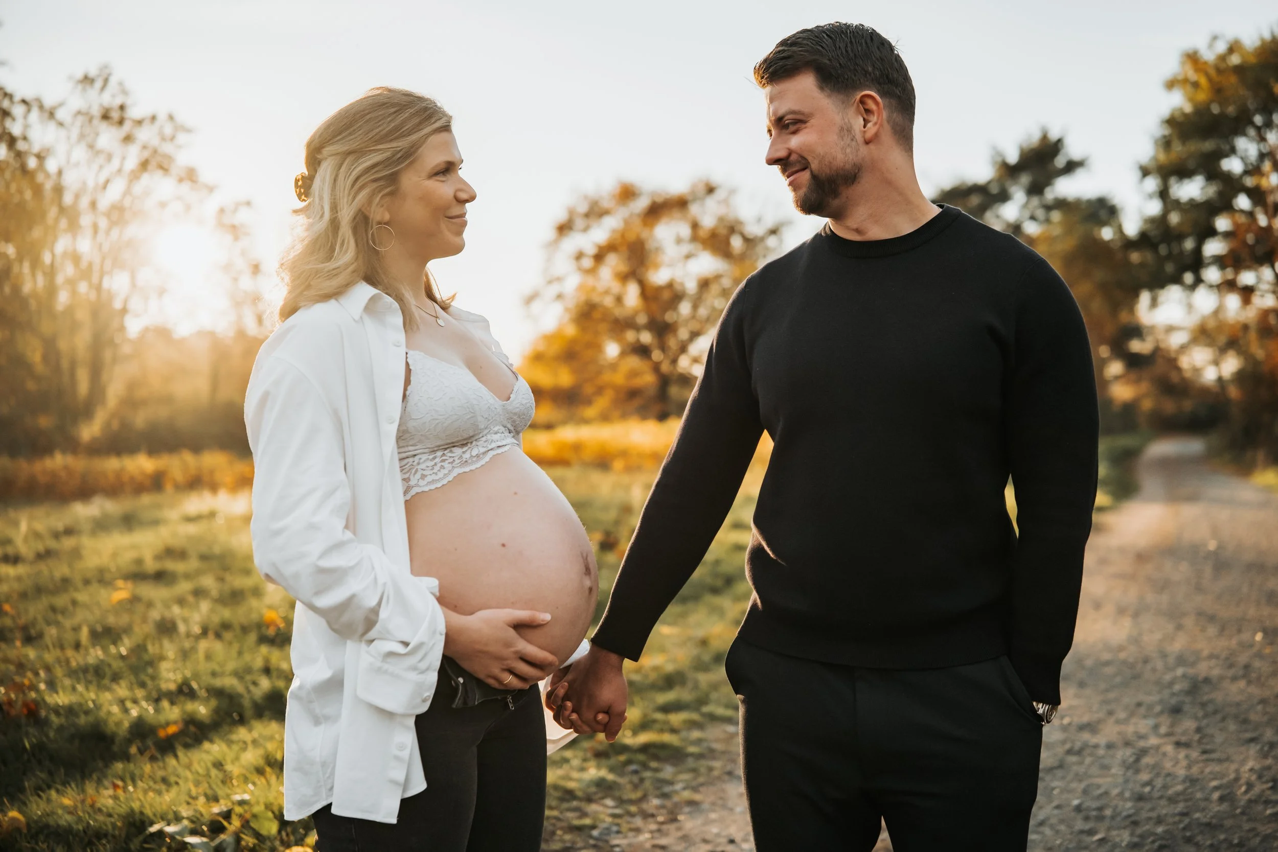 Ein Paar hält Händchen, die schwangere Frau schaut den Mann liebevoll an, im Freien bei Sonnenuntergang, Herbstfarben im Hintergrund.