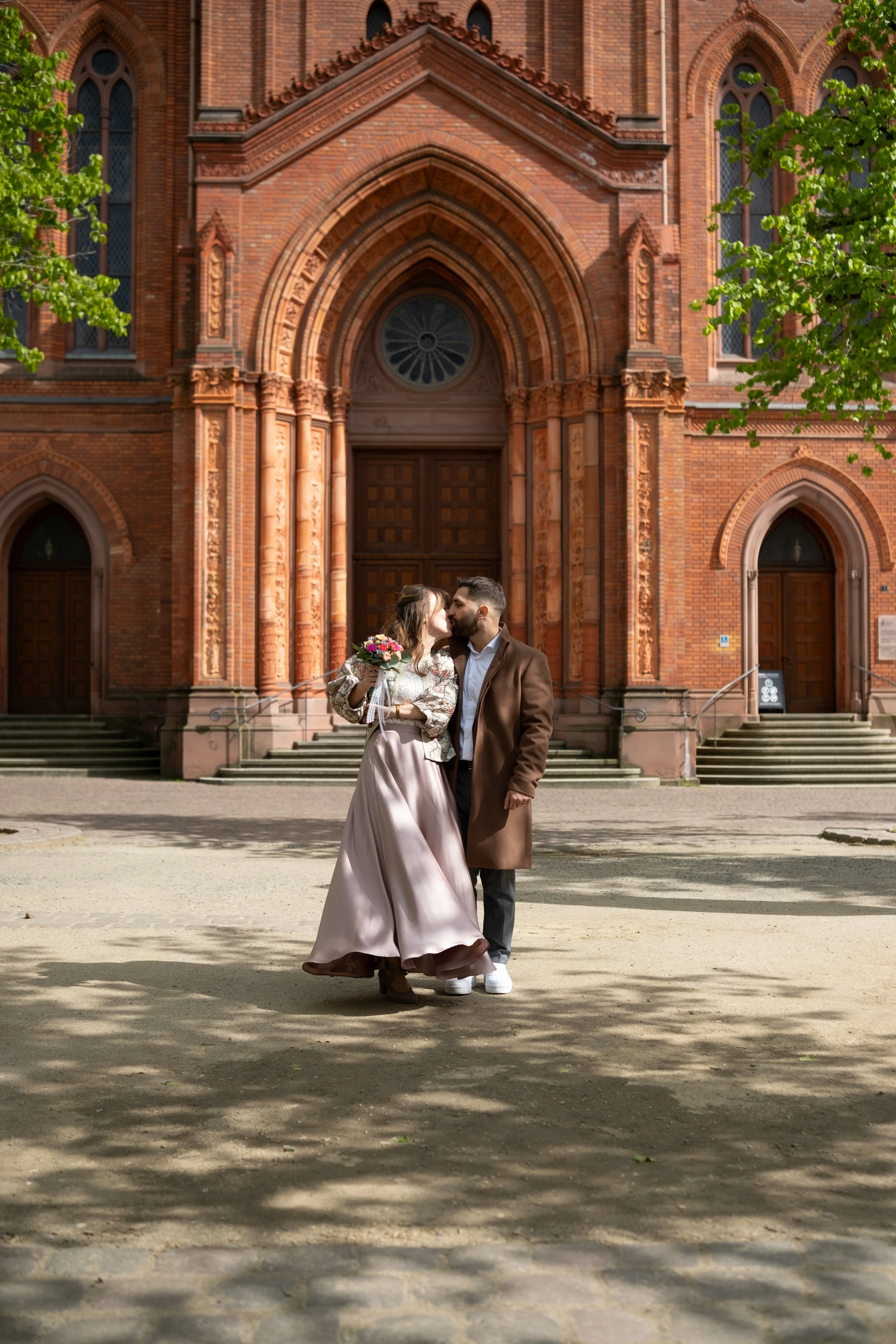 Ein Paar küsst sich vor einer roten Backstein-Kirche, die im Hintergrund steht, während sie eine kleine Brautstrauß hält.