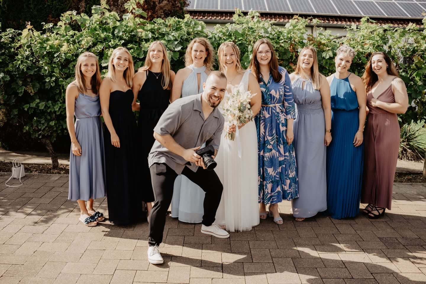 Gruppe von Frauen bei einer Hochzeitsfeier, alle lachen und posieren hinter einem Mann mit Kamera, im Hintergrund grüne Pflanzen.