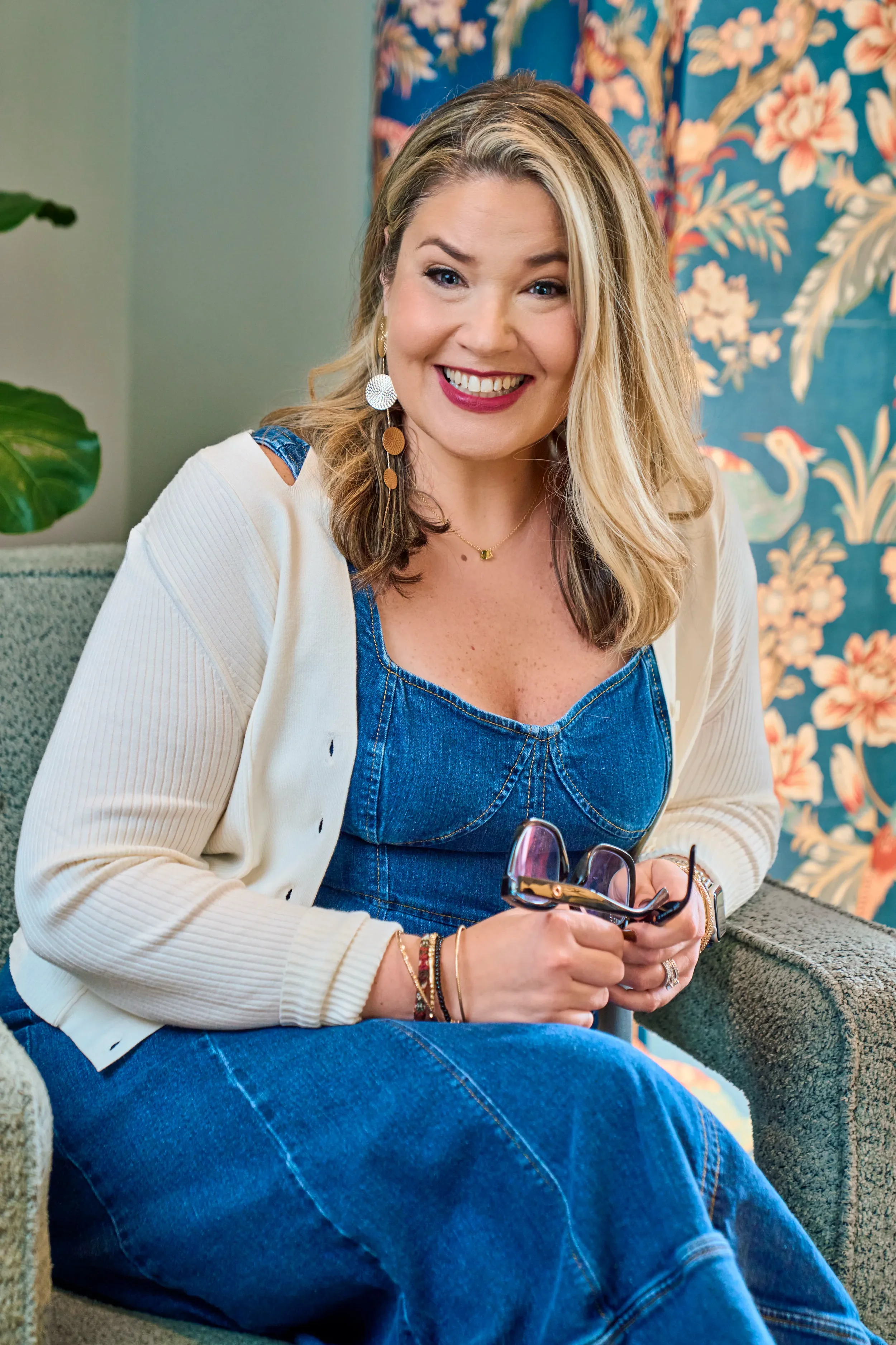 A woman with blonde hair sitting on a grey couch, smiling, holding glasses, wearing a denim top, a beige cardigan, and jewelry, with a floral wallpaper background.