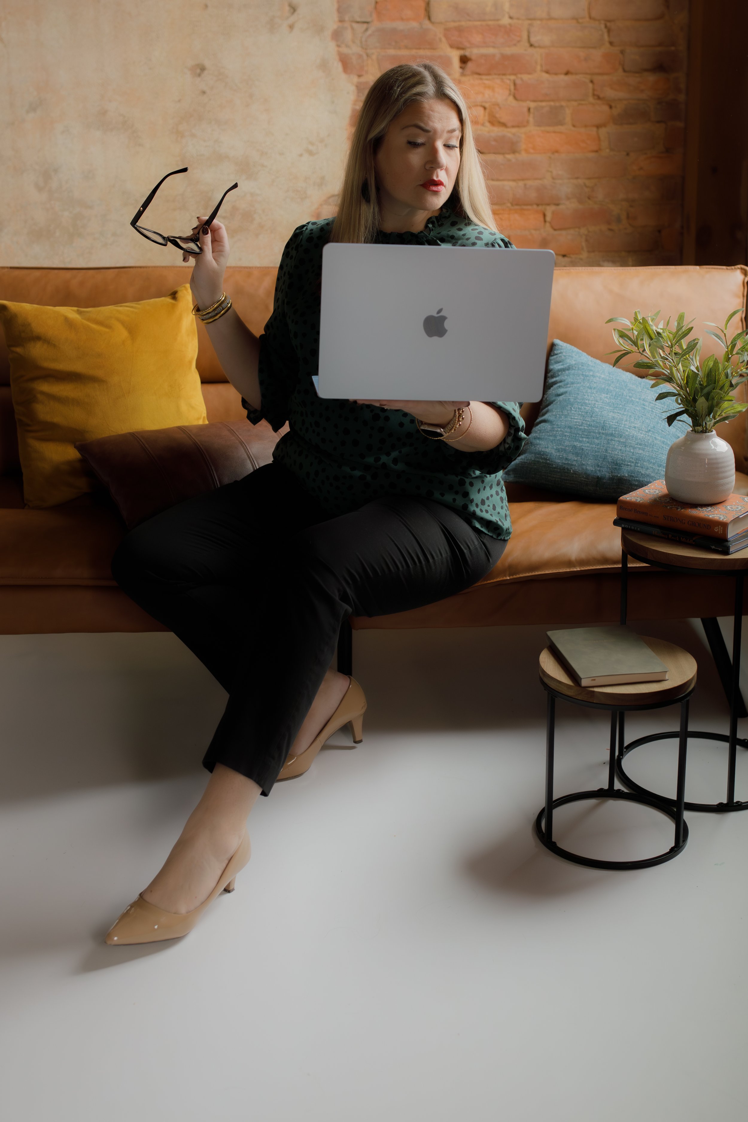 A woman sitting on a leather couch with yellow and blue pillows, holding a pair of glasses in one hand and a silver MacBook in the other, in a room with a brick wall and a small black metal side table with books and a potted plant.