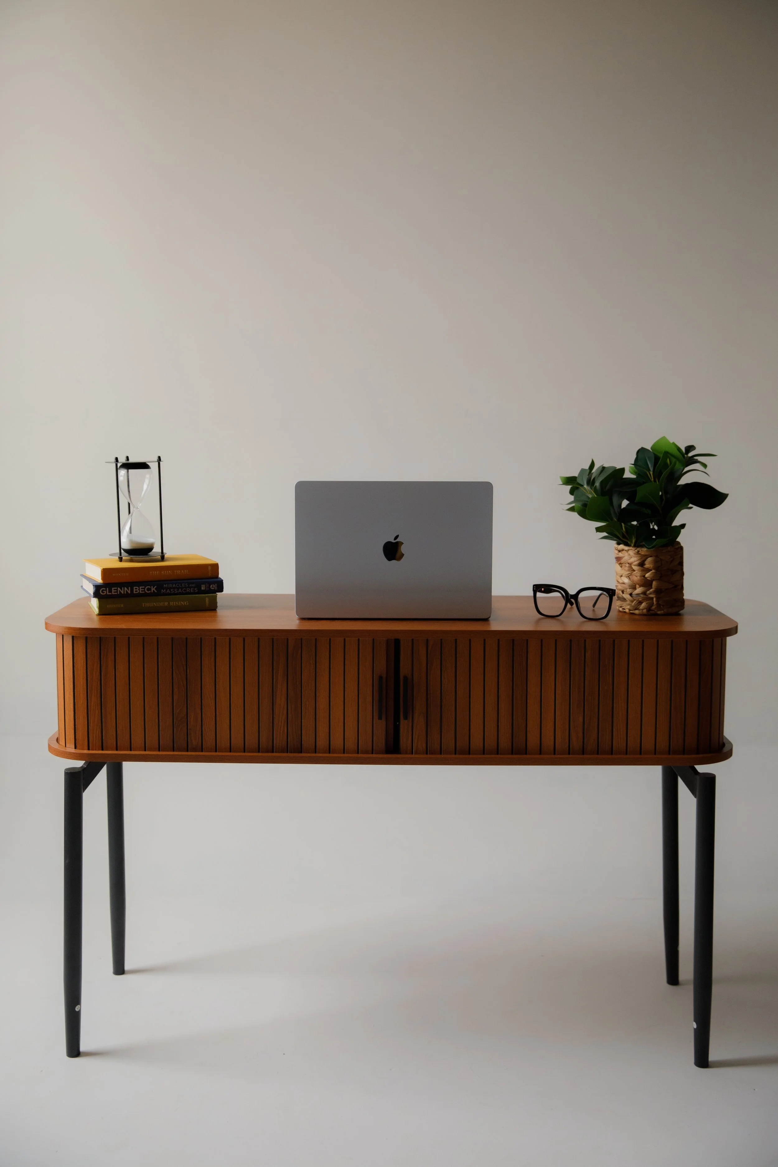 A mid-century modern wooden sideboard with black legs against a plain, light-colored background. On the sideboard, there is an Apple laptop, a pair of black glasses, a potted plant, a stack of books, and an hourglass.