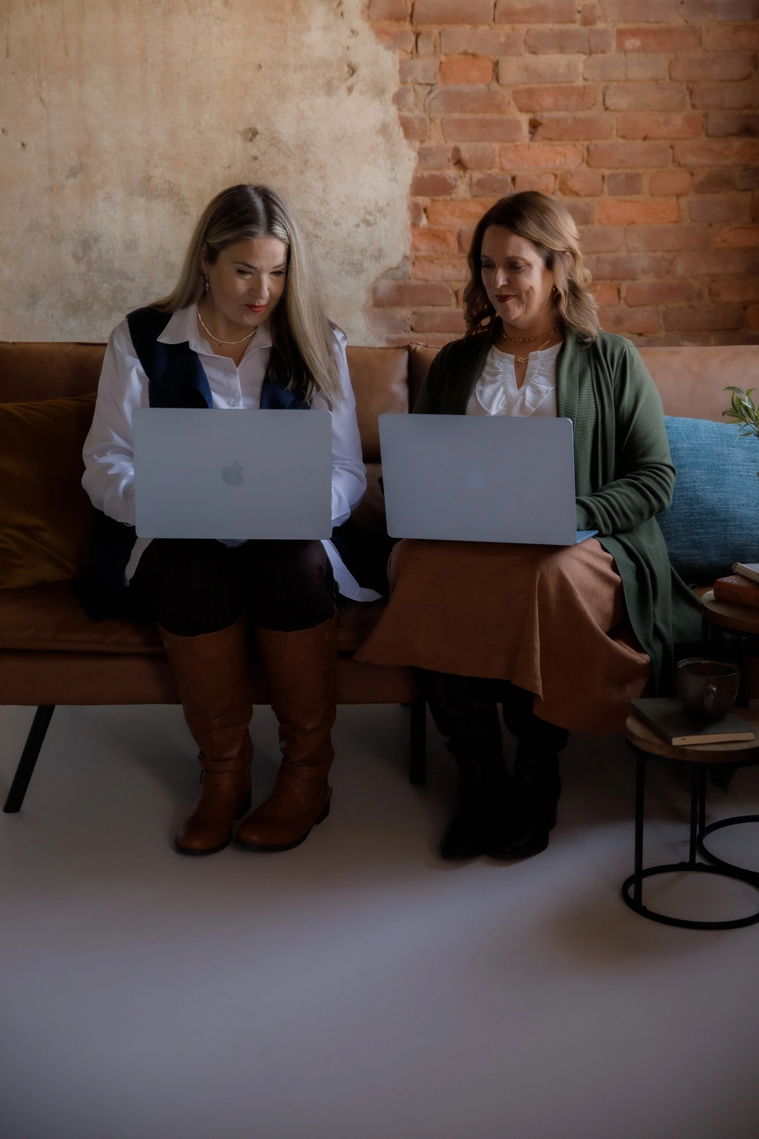 Two women sitting on a brown couch using MacBook laptops, with a brick wall behind them.