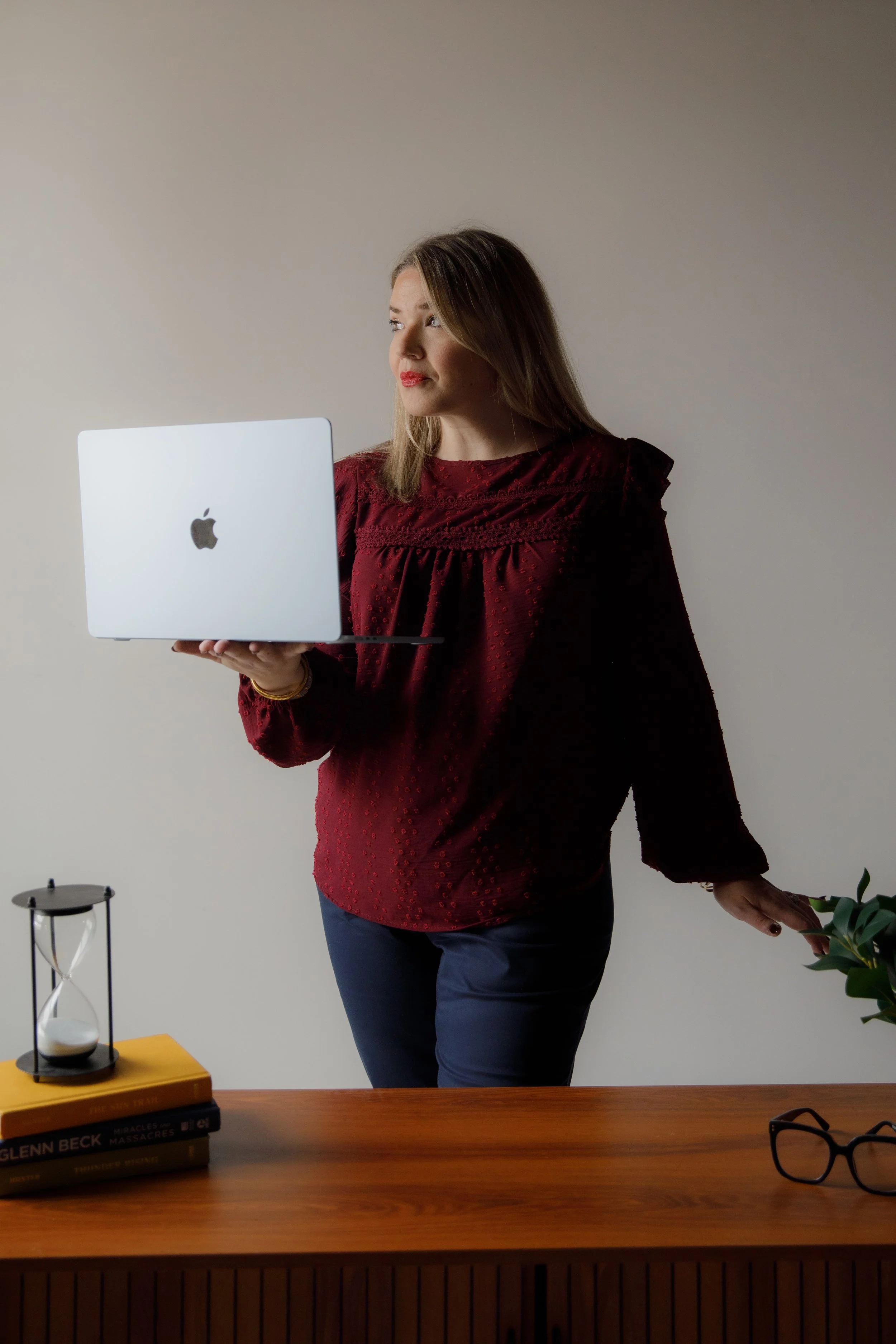 A woman in a red blouse holding a white MacBook in her left hand, standing near a wooden desk with books, glasses, an hourglass, and a plant. She is looking to her left.