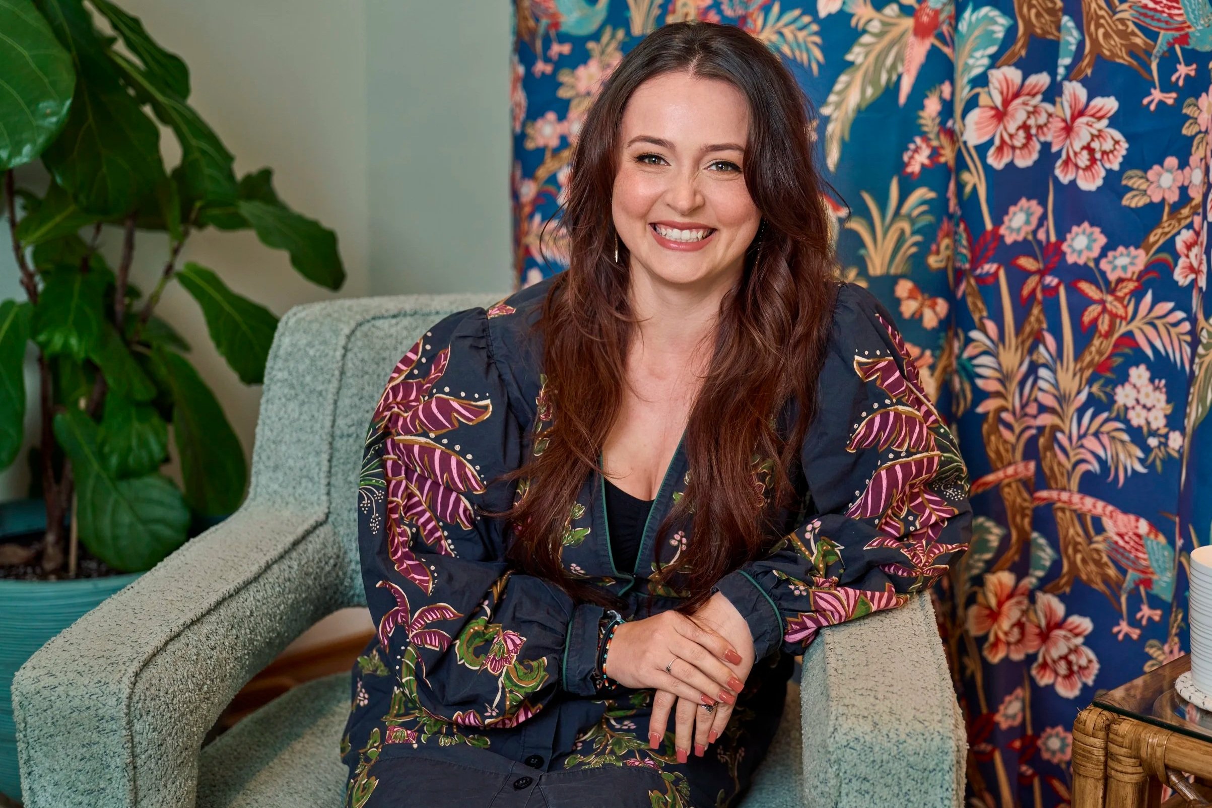 A woman with long brown hair, smiling, sitting on a light green textured sofa, wearing a navy jacket with colorful floral patterns. Behind her, a vibrant floral patterned fabric or wall hanging. To her left, there's a large leafy green houseplant, and on her right, stacked white cups on a wooden table.