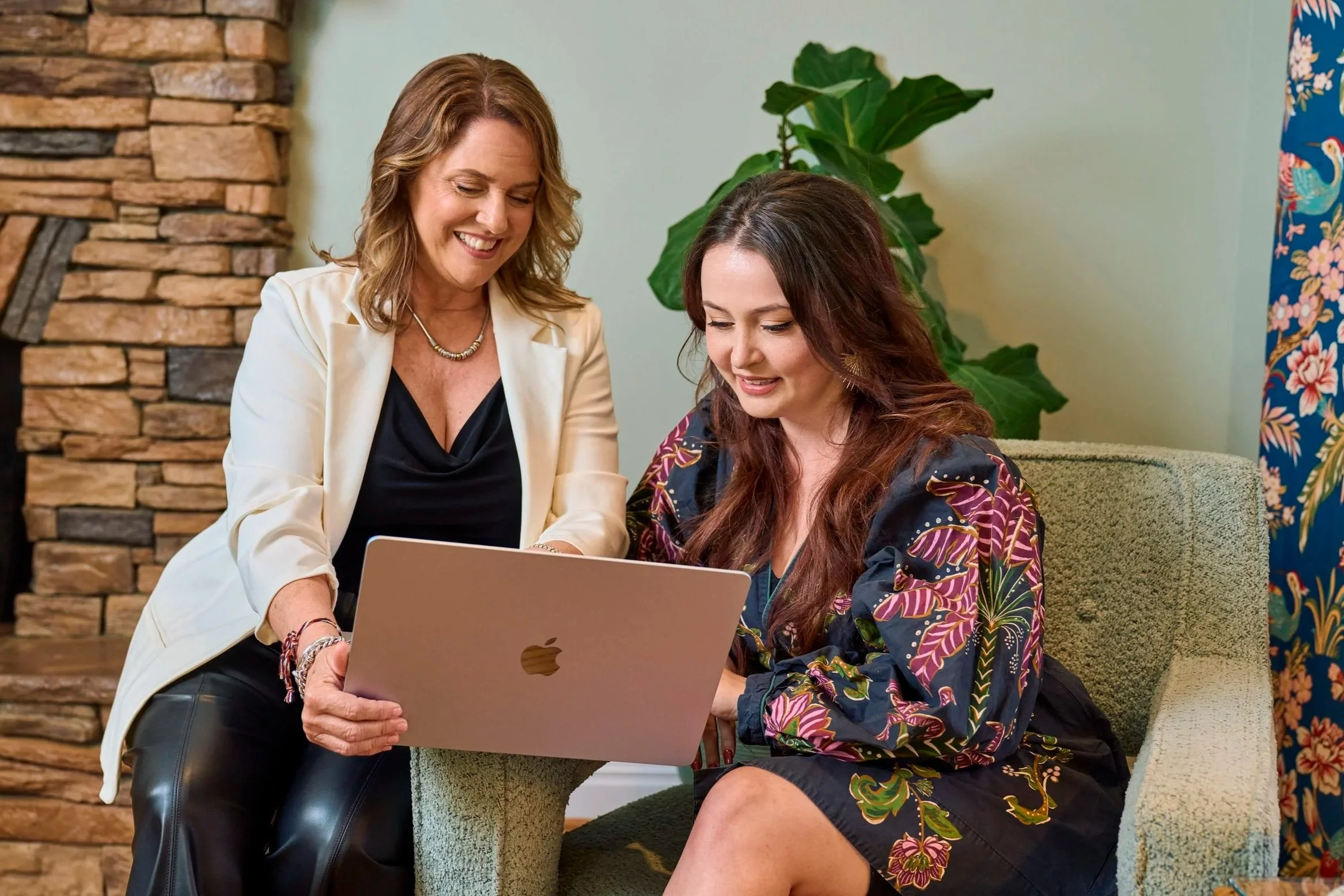 Two women sitting on a chair, looking at a laptop. One woman is standing, wearing a white blazer and black top, smiling. The other woman is sitting, wearing a colorful, patterned top, also smiling. Background includes a stone fireplace, green plant, and floral curtain.