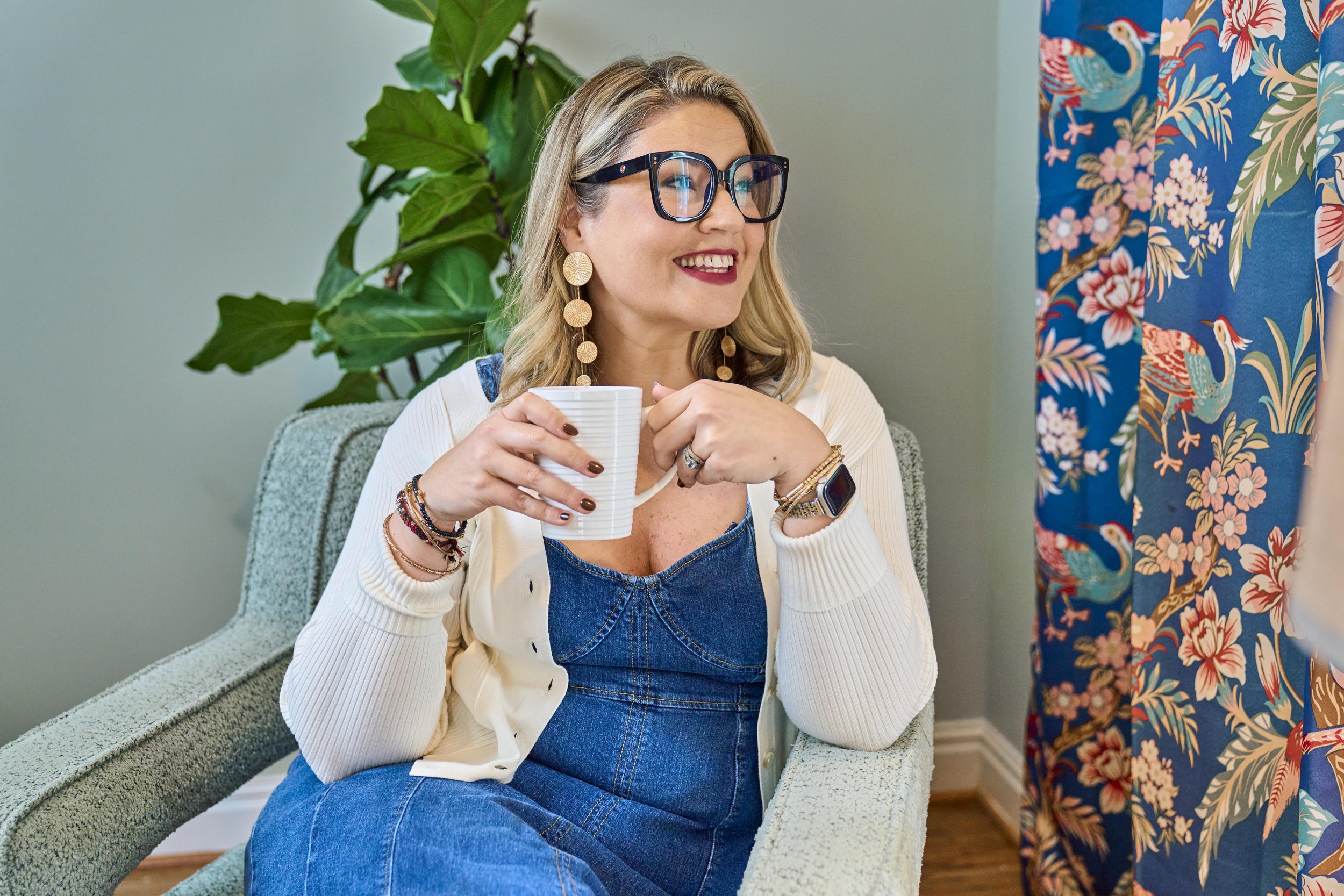 A woman with blonde hair, wearing large black glasses, gold earrings, a white cardigan, a denim top, and multiple bracelets, smiling while sitting on a marble bench holding a white mug. She is in a room with green plants and decorative blue curtains with a colorful bird and floral pattern.