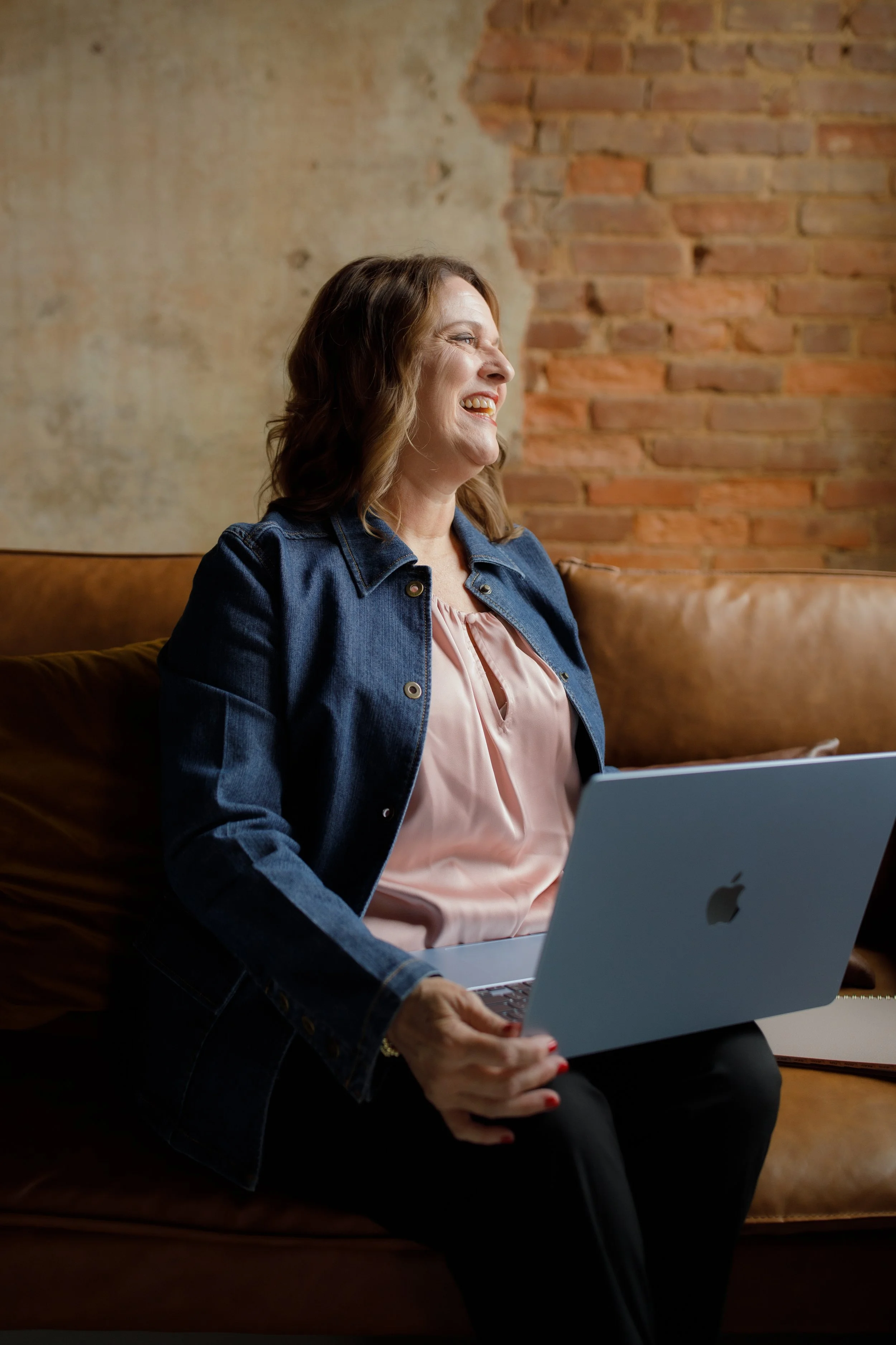A woman sitting on a brown leather couch, holding a silver Apple MacBook laptop, smiling and looking to her left, with a brick wall and textured ceiling in the background.