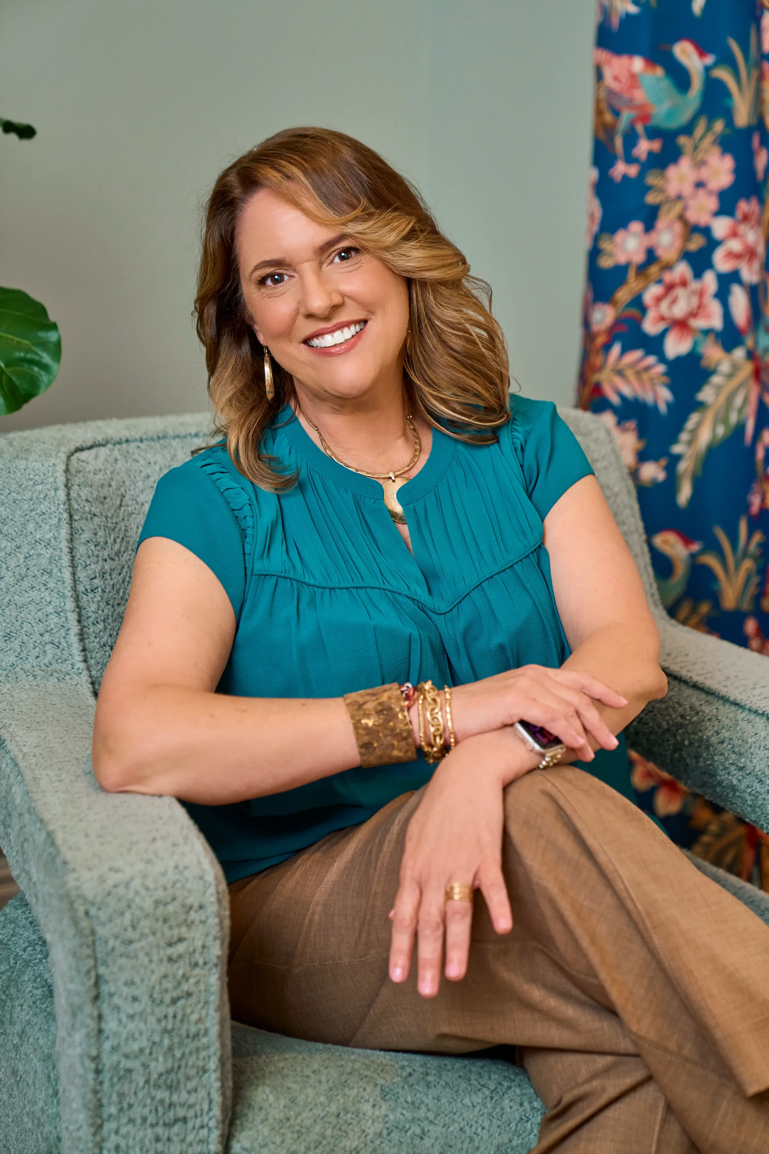 A woman with light brown hair and a bright smile sitting on a light green sofa, wearing a teal blouse and brown pants, accessorized with gold jewelry, in a room with a plant and a floral curtain.