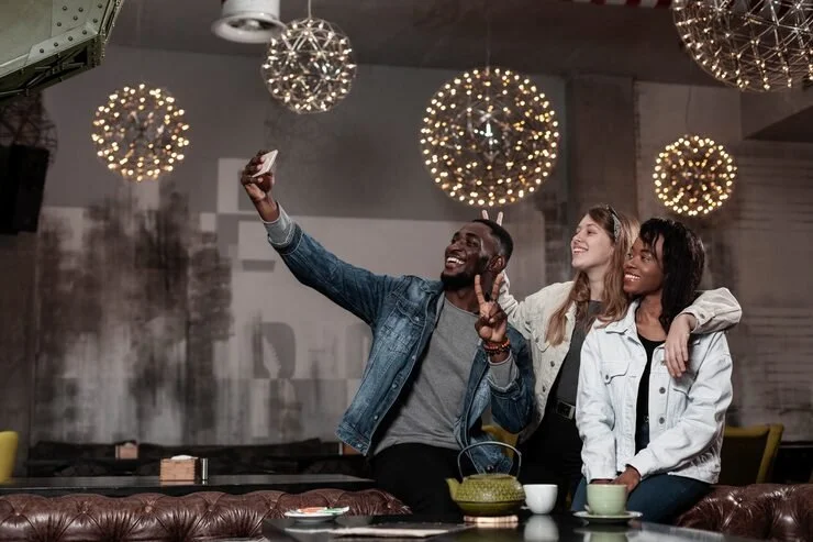 Three friends taking a selfie together at a social gathering, smiling and posing in a cozy lounge with hanging spherical lights and a modern decor.