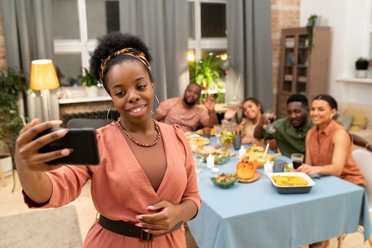A young woman takes a selfie with friends at a dinner party in a warmly lit living room. Four friends are seated at a table with food, smiling and waving at the camera.