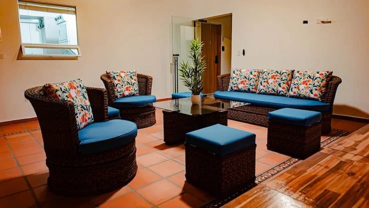 Living room with wicker furniture, blue cushions, and floral patterned pillows, a glass coffee table with a potted plant, terracotta tile floor, and a window showing an indoor space.