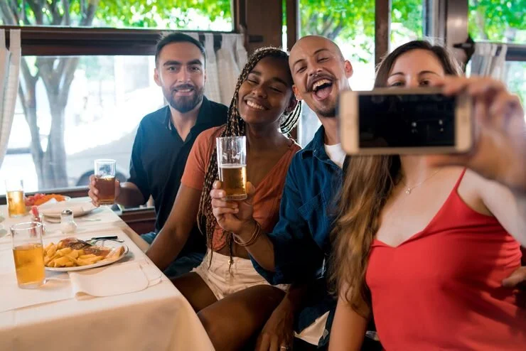 Four friends sitting at a restaurant table, smiling and taking a selfie. The table has food and drinks, and there are large windows with greenery outside in the background.