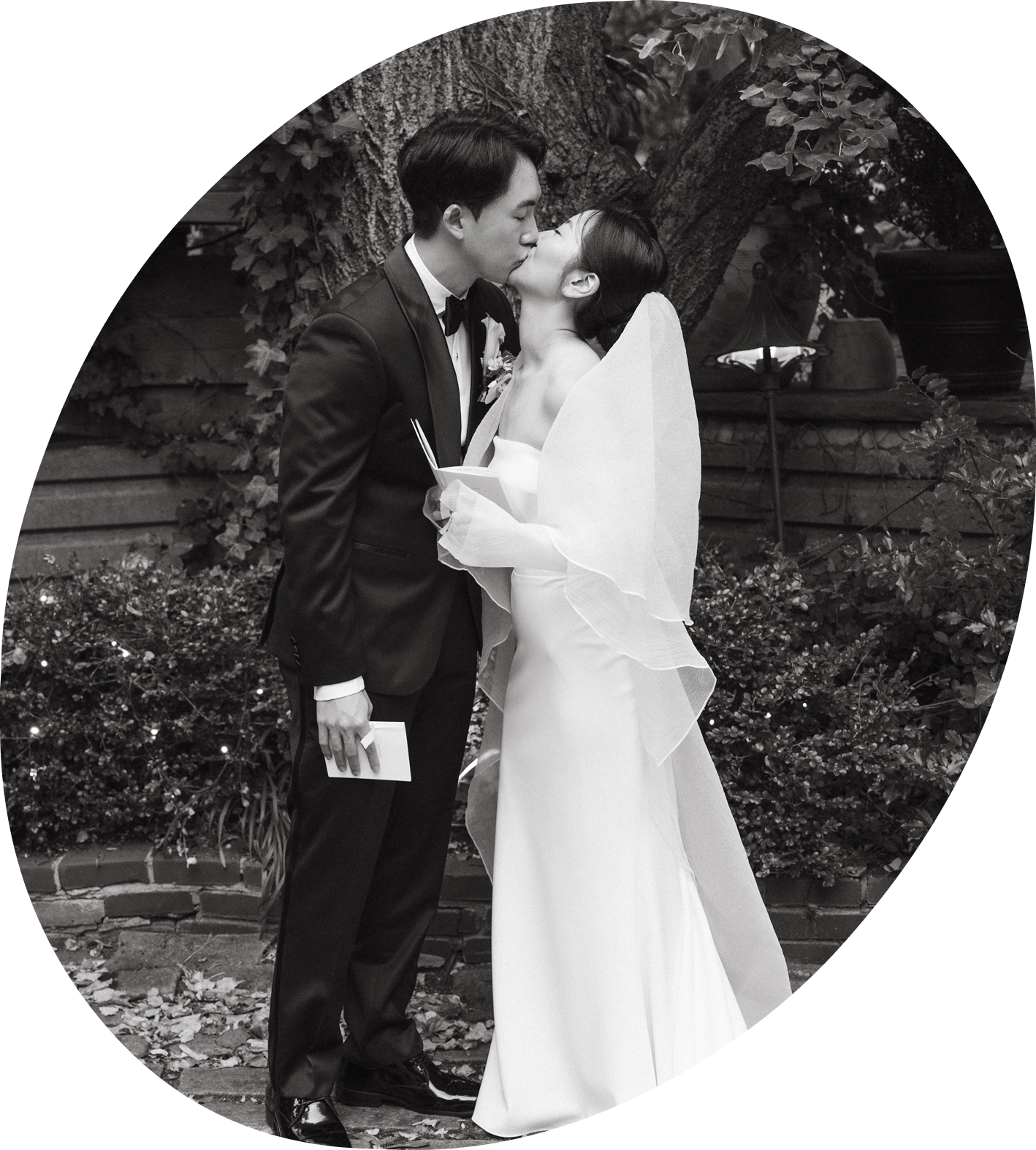 A black-and-white photo of a bride and groom kissing outdoors during their wedding ceremony. The groom is in a tuxedo holding a small book, and the bride is in a wedding dress with a sheer veil. They stand in front of a large tree and some steps with potted plants in the background.