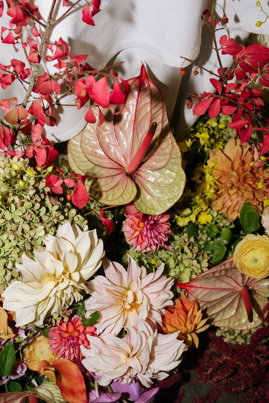 An assortment of colorful flowers and leaves, including white, pink, and yellow dahlias, red and pink anthuriums, red and pink bougainvillea, yellow yarrow, and various green foliage.