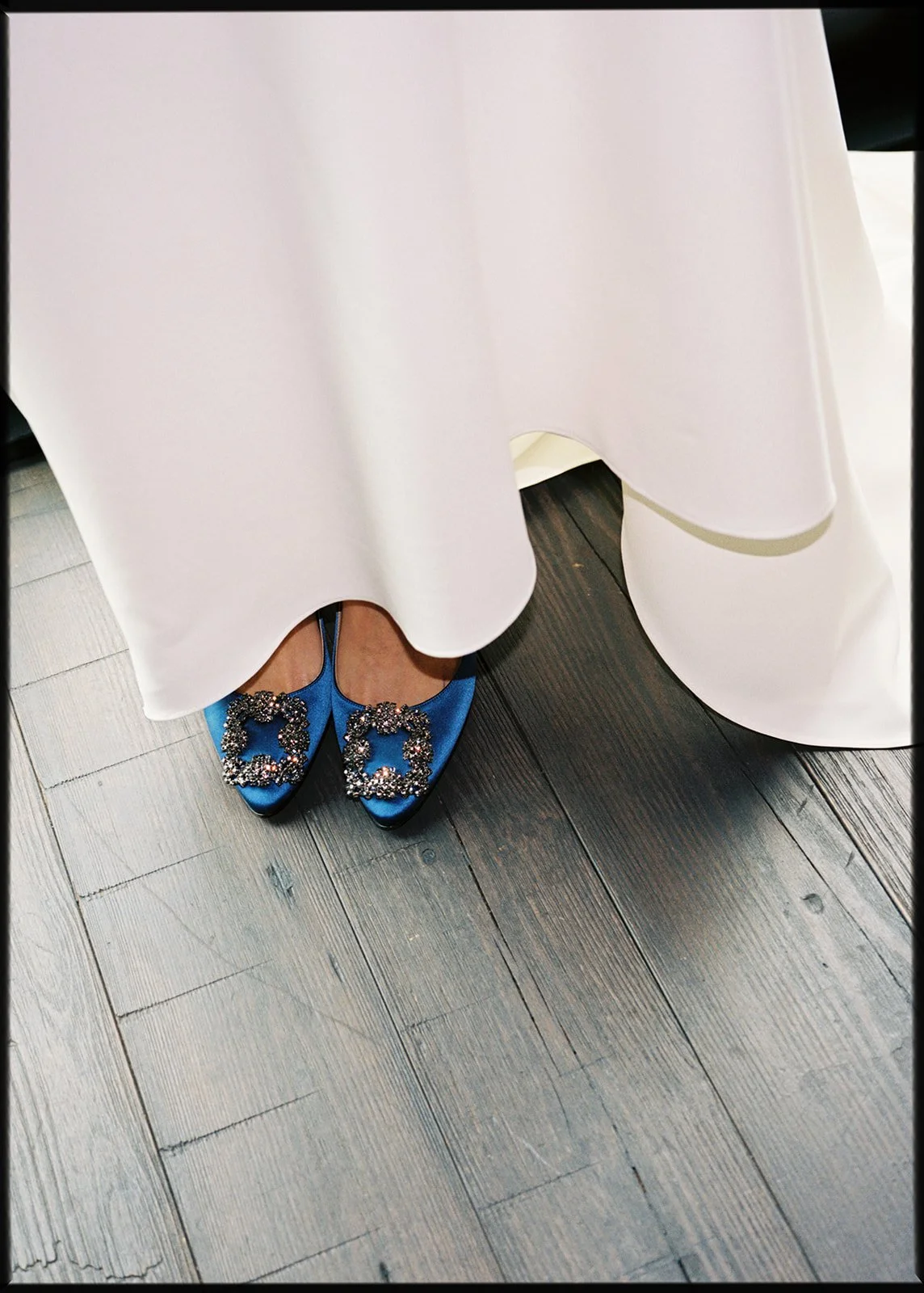 Close-up of feet wearing blue satin shoes with large rhinestone bows, partially covered by an elegant white dress, standing on dark wooden floor.