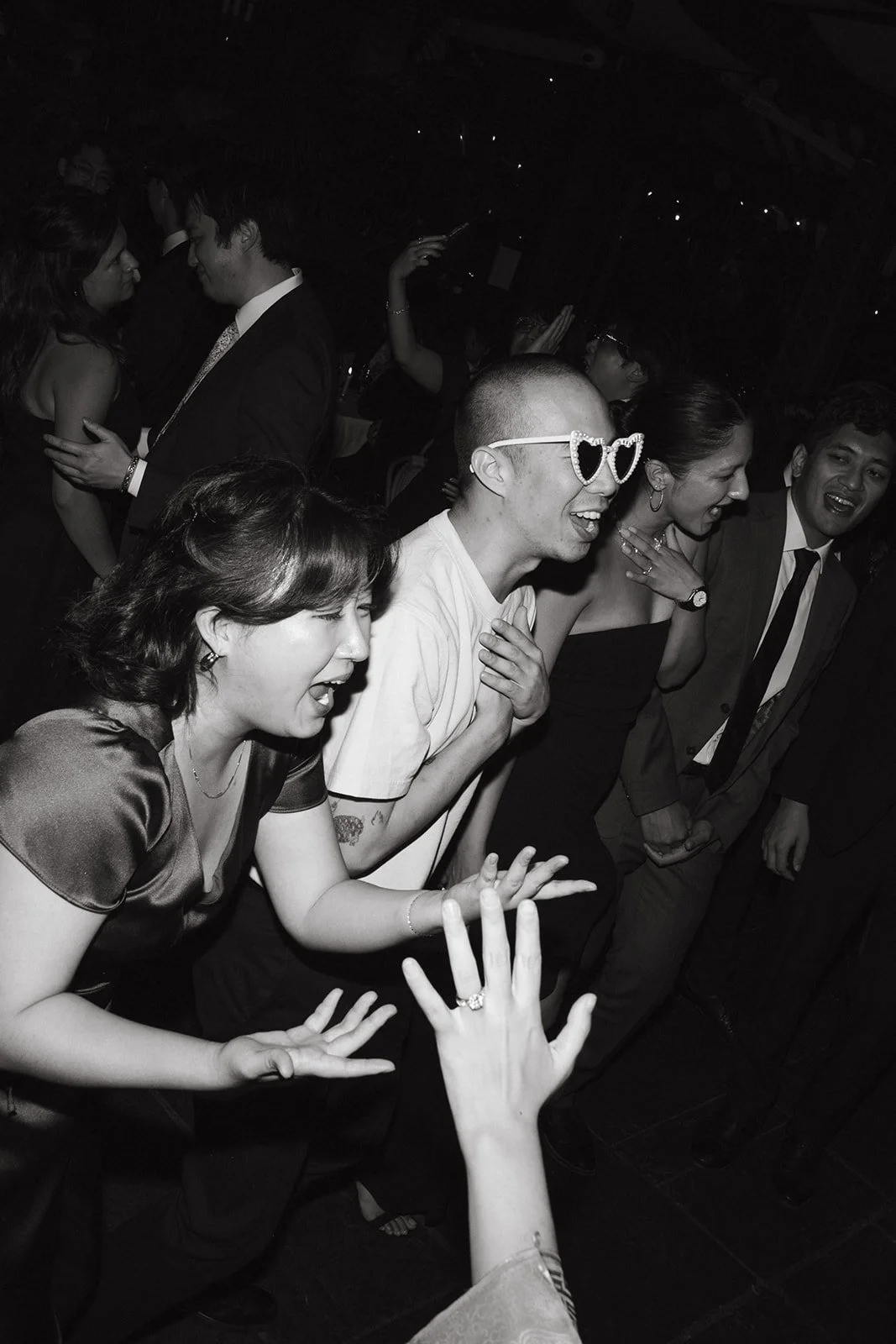 People dancing and celebrating at a party, some wearing sunglasses and formal attire, with smiling and laughing faces.