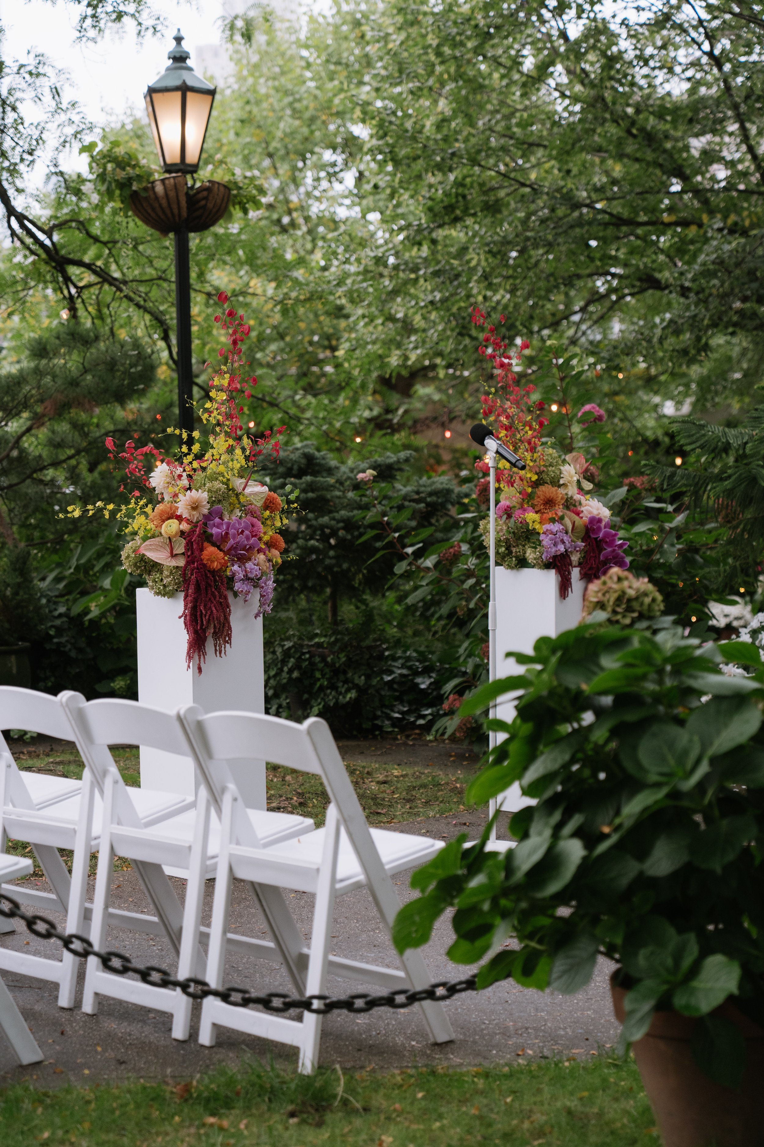 Empty white chairs arranged in rows in an outdoor garden setting, decorated with floral arrangements on tall white pedestals, with a vintage lamppost and greenery in the background.