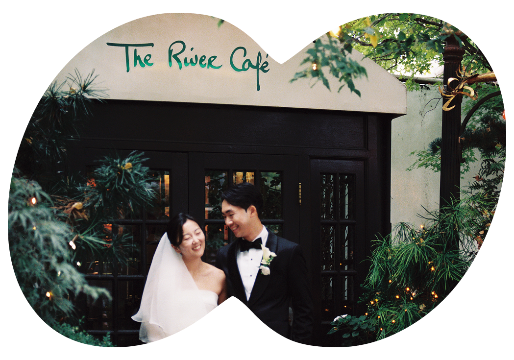 A bride and groom standing close together outdoors in front of a building with a sign that reads "The Riverside Cafe." The bride is wearing a white wedding dress and veil, and the groom is dressed in a black tuxedo with a bow tie. They are smiling and looking at each other, surrounded by greenery and decorative lights.