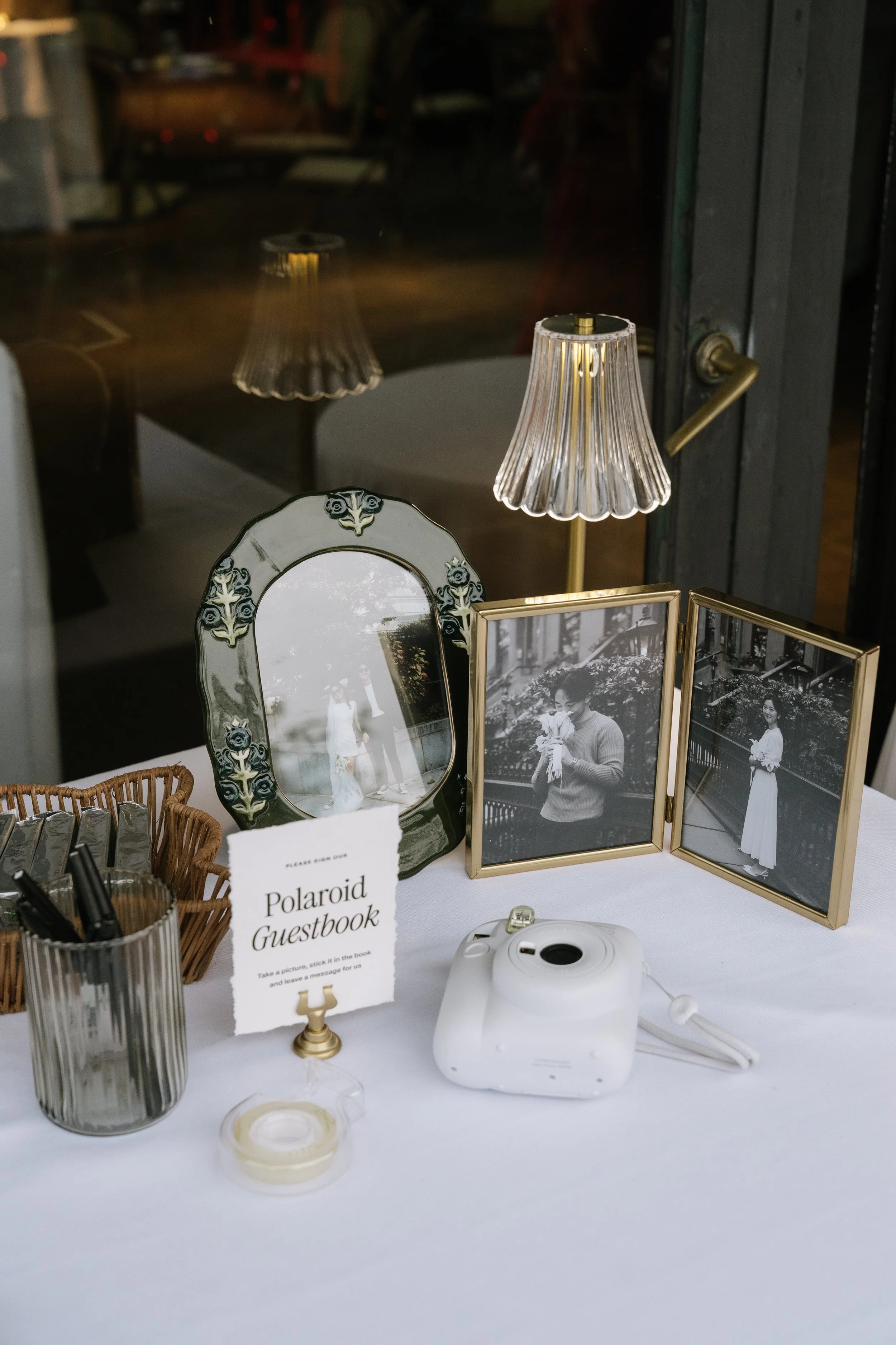 A Polaroid guestbook table featuring framed black-and-white wedding photos, a small mirror, candles, pens, hand sanitizer, a Polaroid camera, and a sign instructing guests to take a picture and leave a message.