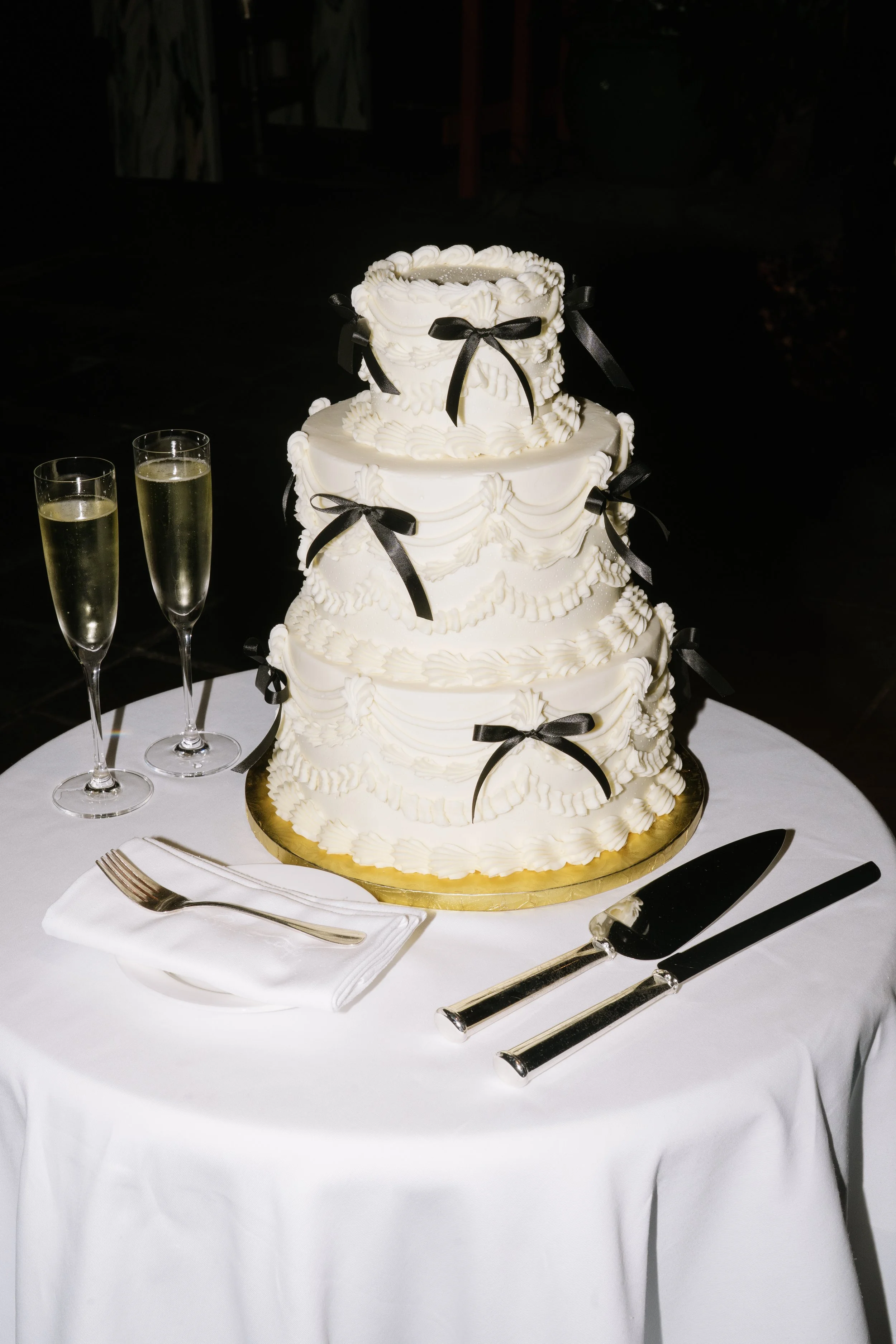 A four-tiered white wedding cake decorated with black ribbons, placed on a gold cake board. The cake is on a round table with a white tablecloth, set with a cake knife, server, fork, a napkin, and two glasses of champagne.