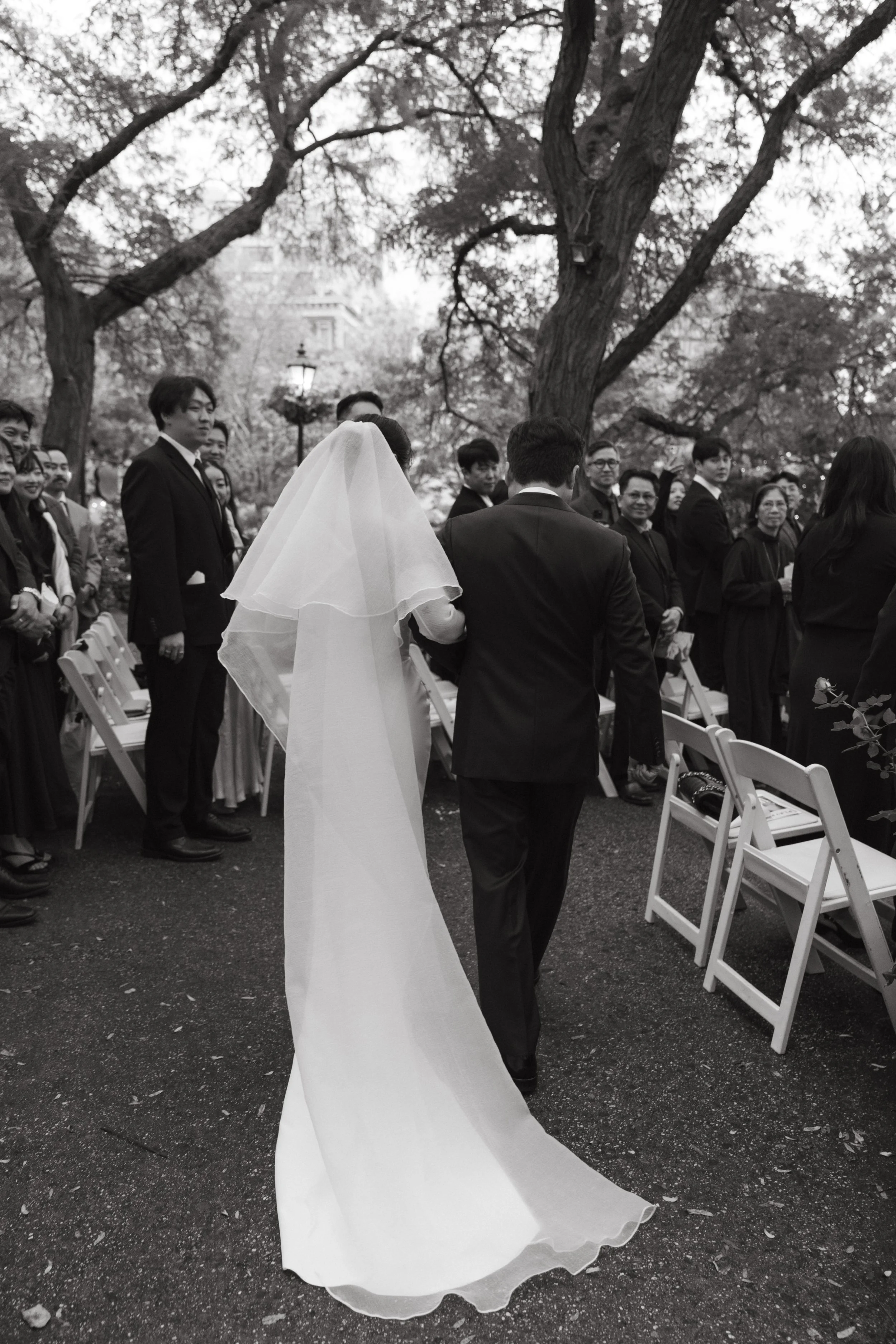 A black-and-white photo of a wedding ceremony outdoors, showing a bride in a veil and gown walking arm-in-arm with a groom in a suit, surrounded by seated guests and tall trees.