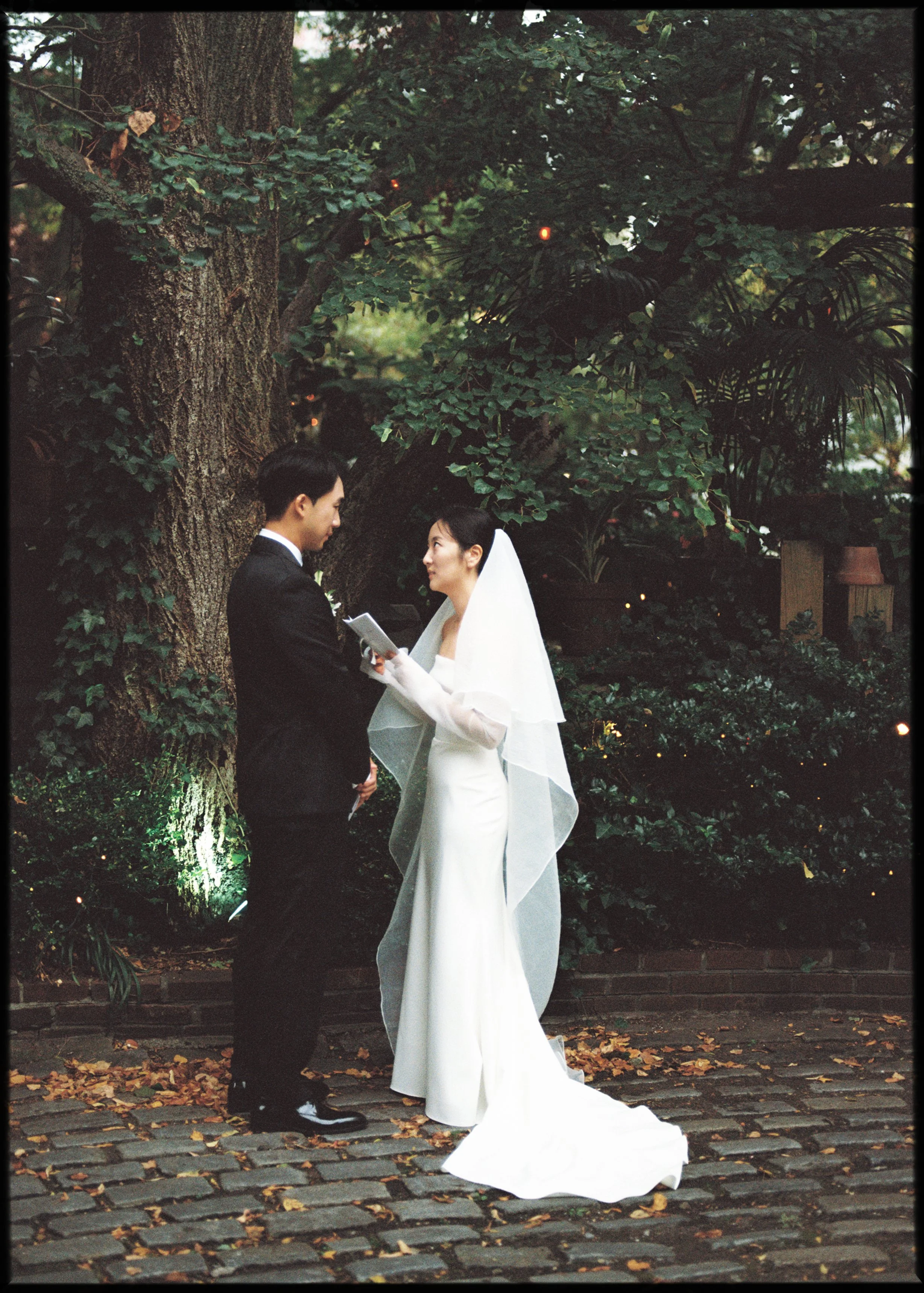A bride and groom standing outdoors during their wedding ceremony, exchanging vows under a large tree surrounded by greenery, with fallen leaves on the ground.
