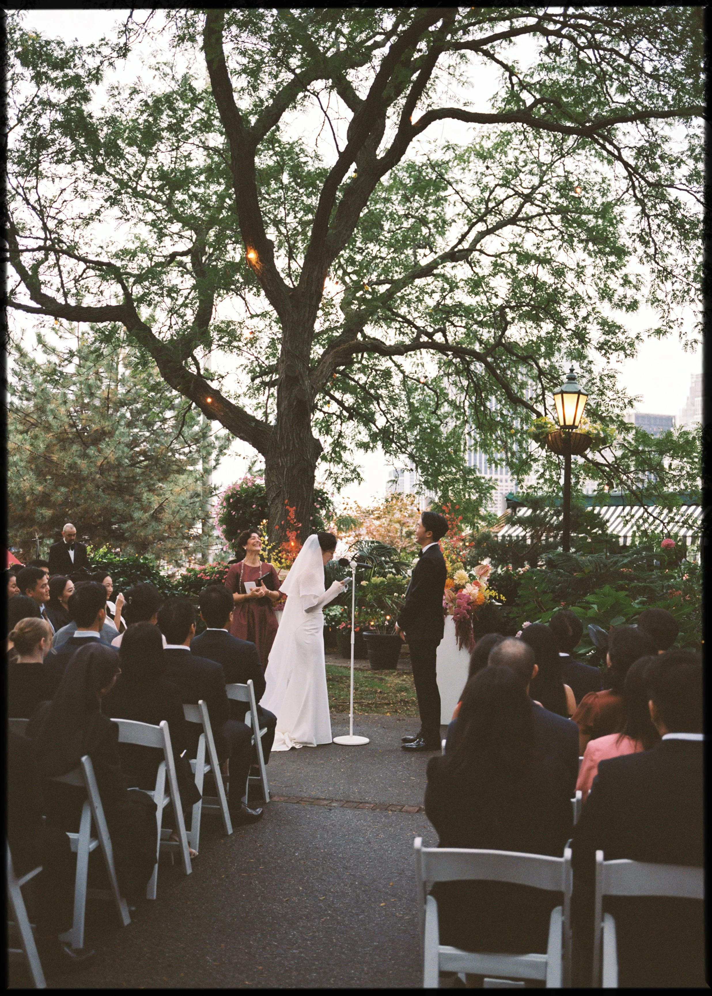 A wedding ceremony taking place outdoors under a large tree with green leaves, with a bride in a white dress and veil and a groom in a black suit facing each other. The officiant and guests are seated around them. The setting is decorated with flower