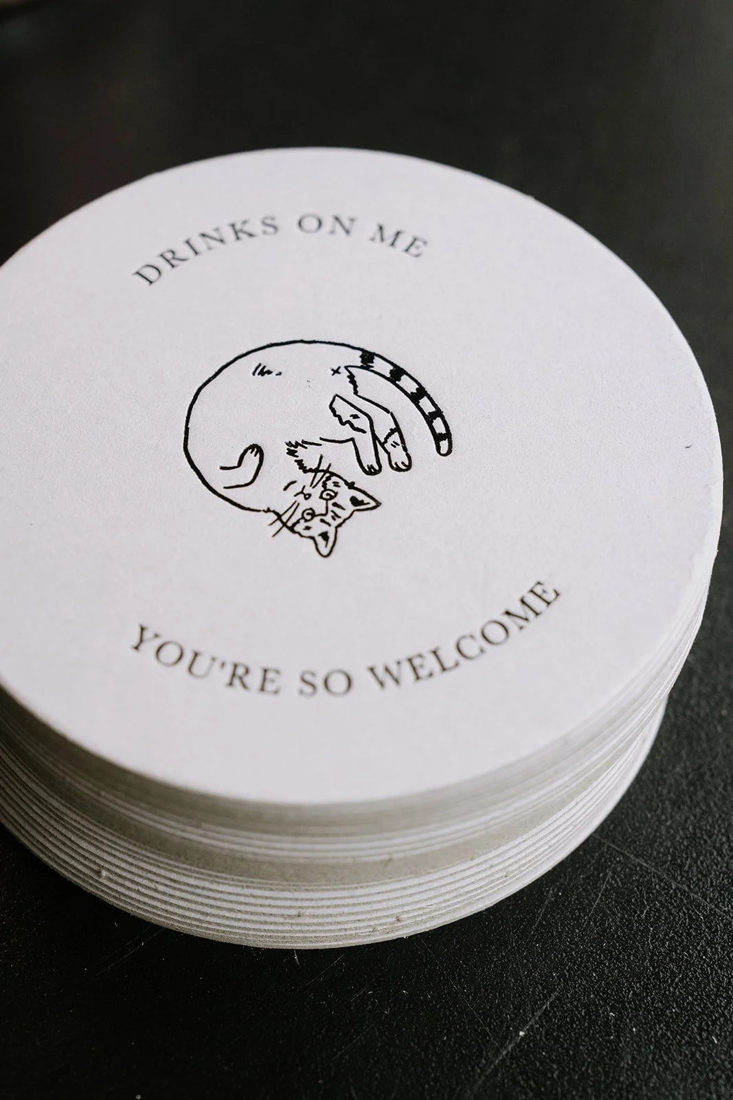 Stack of coasters with a printed illustration of a cat curled up and the text 'Drinks on me, you're so welcome' surrounding it.