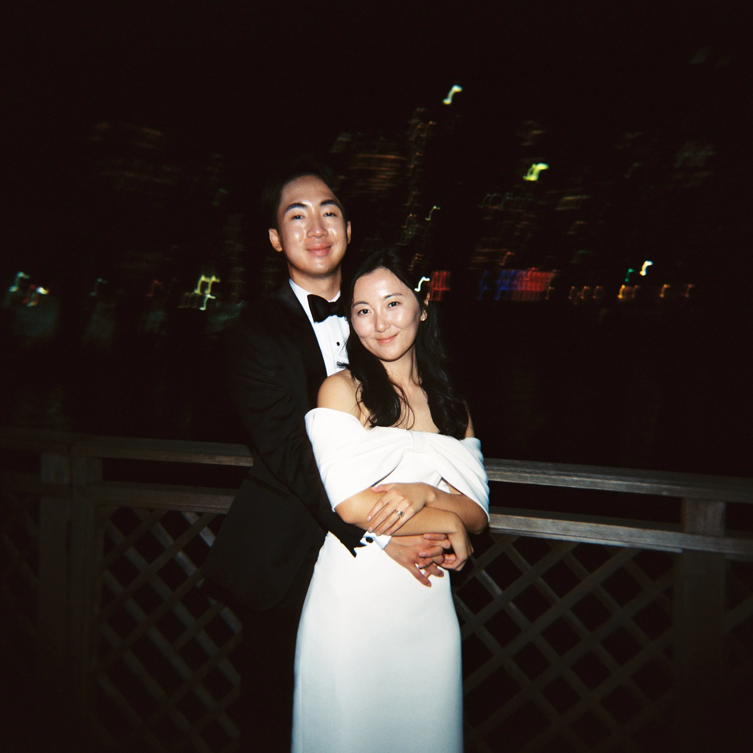 A couple dressed in formal attire at night, standing on a balcony with city lights in the background, smiling and embracing each other.