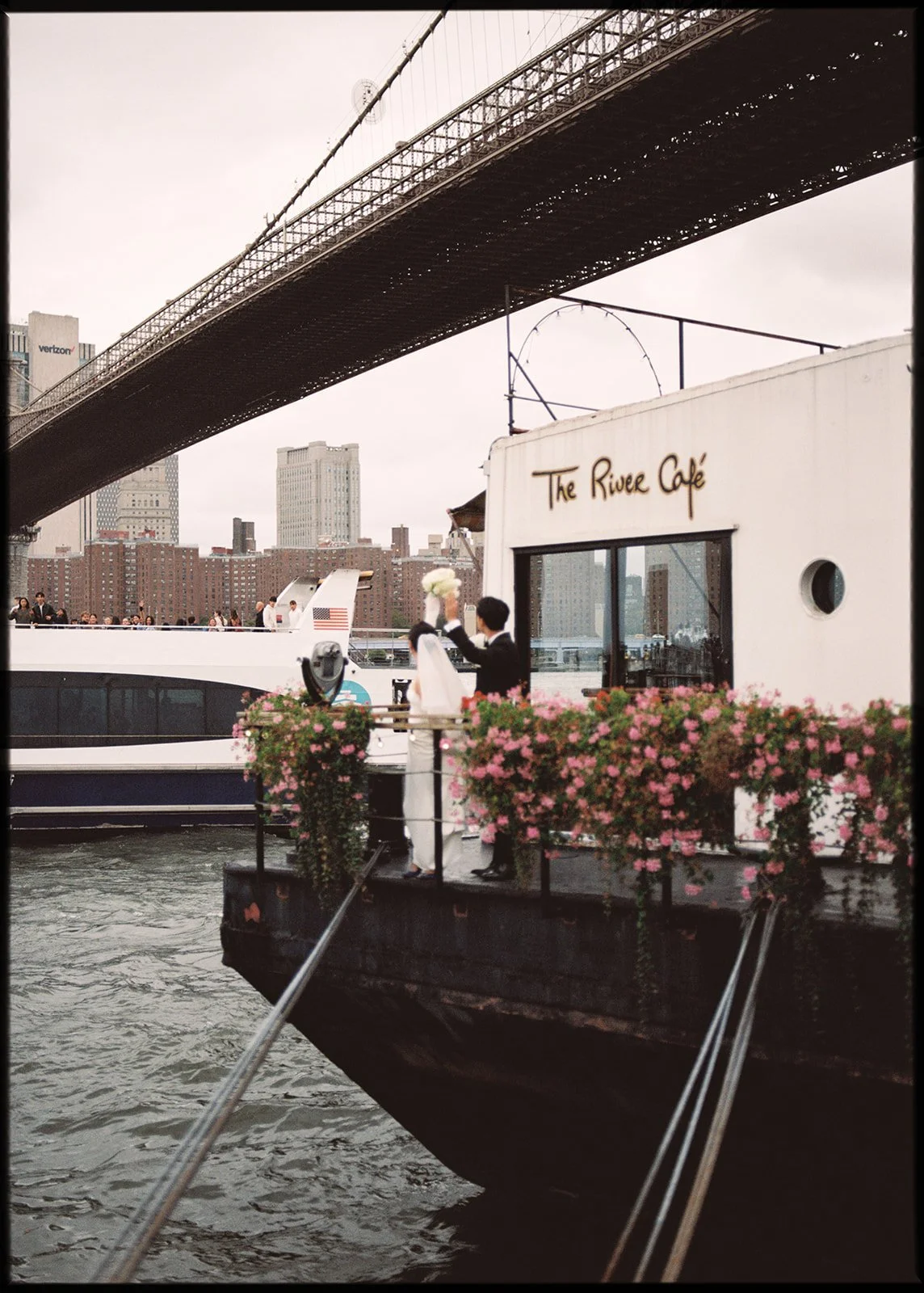 A couple in wedding attire on a boat named The River Café, decorated with pink flowers, under a bridge in New York City, with a city skyline in the background.