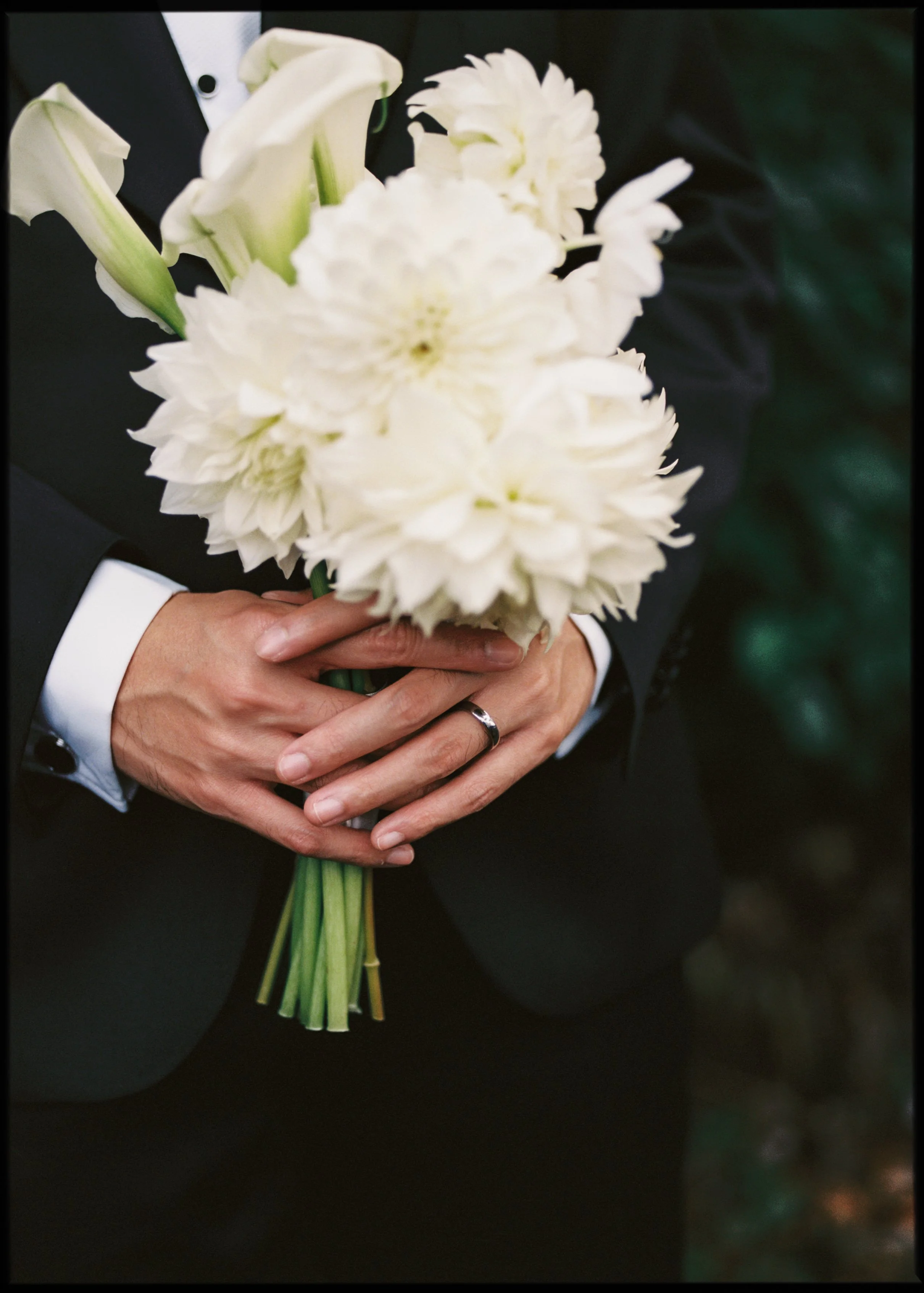 Person in a dark suit holding a bouquet of white lilies and dahlias with both hands, wearing a wedding or engagement ring.