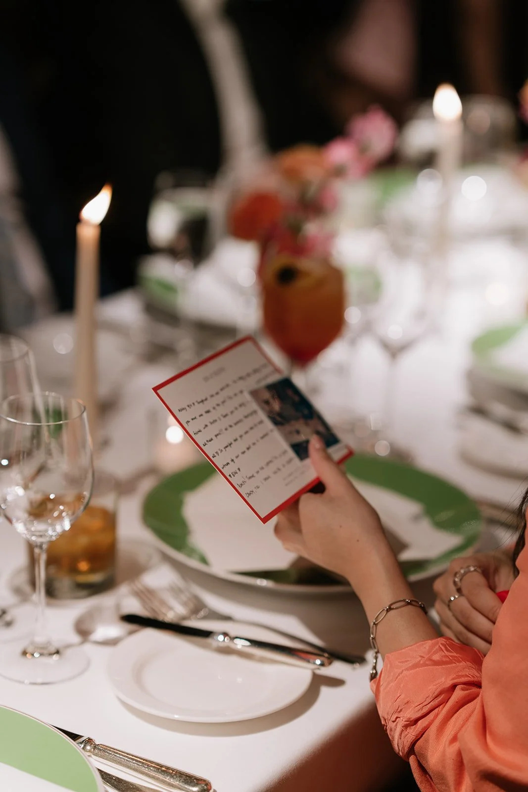 A person holding a red-bordered card over a white and green dinner plate at a formal event with candles and floral decorations on a decorated table.