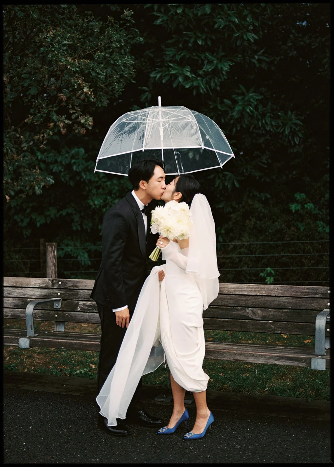 A newlywed couple sharing a kiss under a transparent umbrella on a rainy day, standing in front of a wooden bench and lush green trees. The groom is in a black suit, and the bride is in a white dress holding a bouquet of white flowers, wearing blue h