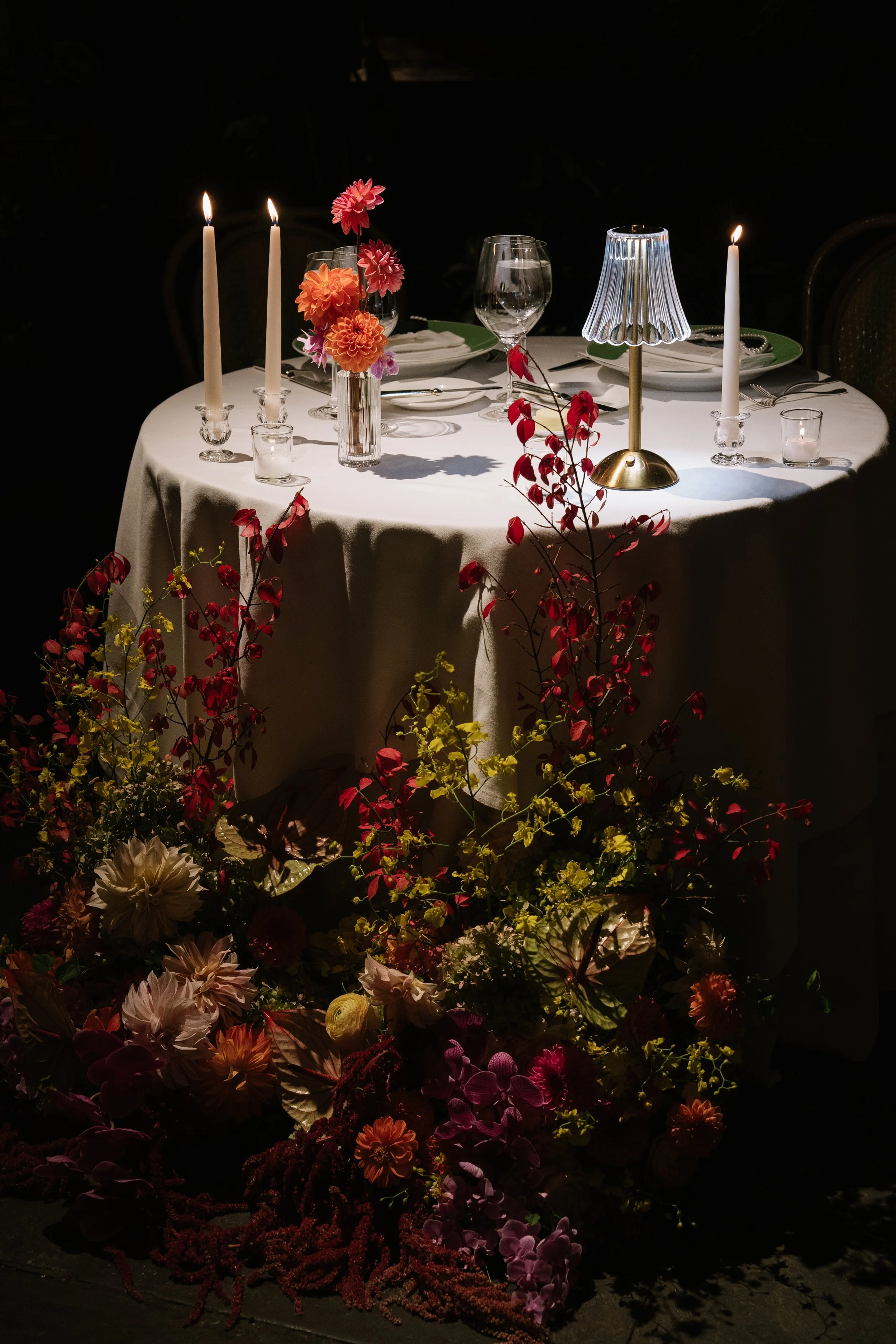 A table set for a dinner with candles, glasses, and flowers, illuminated in a dark setting, with an arrangement of colorful flowers at the base.