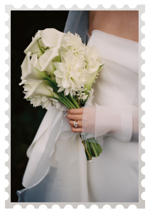 Close-up of a bride holding a bouquet of white flowers, including roses and chrysanthemums, with a wedding ring visible on her finger.