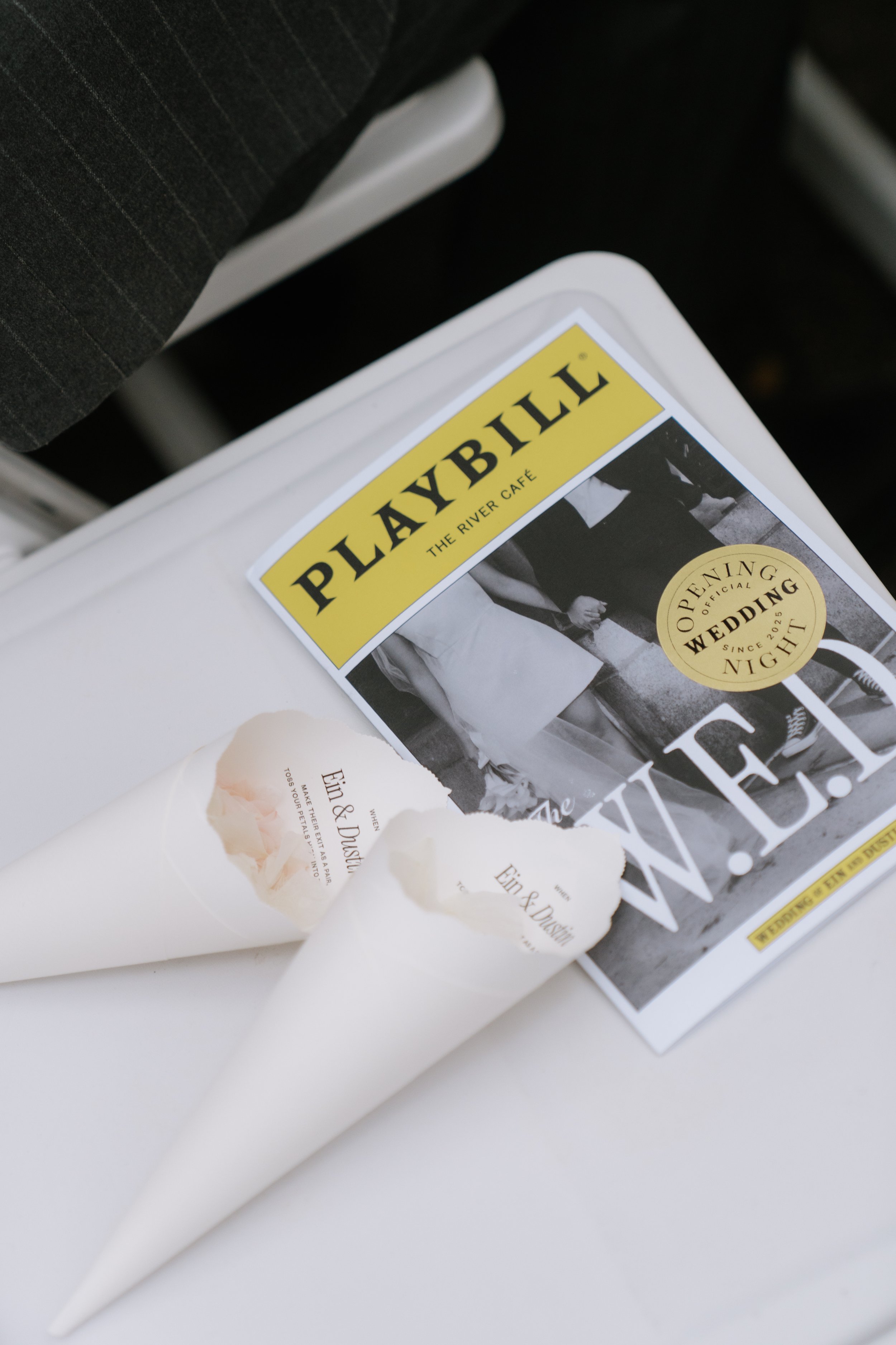 A wedding invitation and two cone-shaped paper fans on a white surface. The invitation is for a wedding at Playbill, The River Café, with a black and white photo of a couple and a gold circle with black text about the opening of the wedding night sin