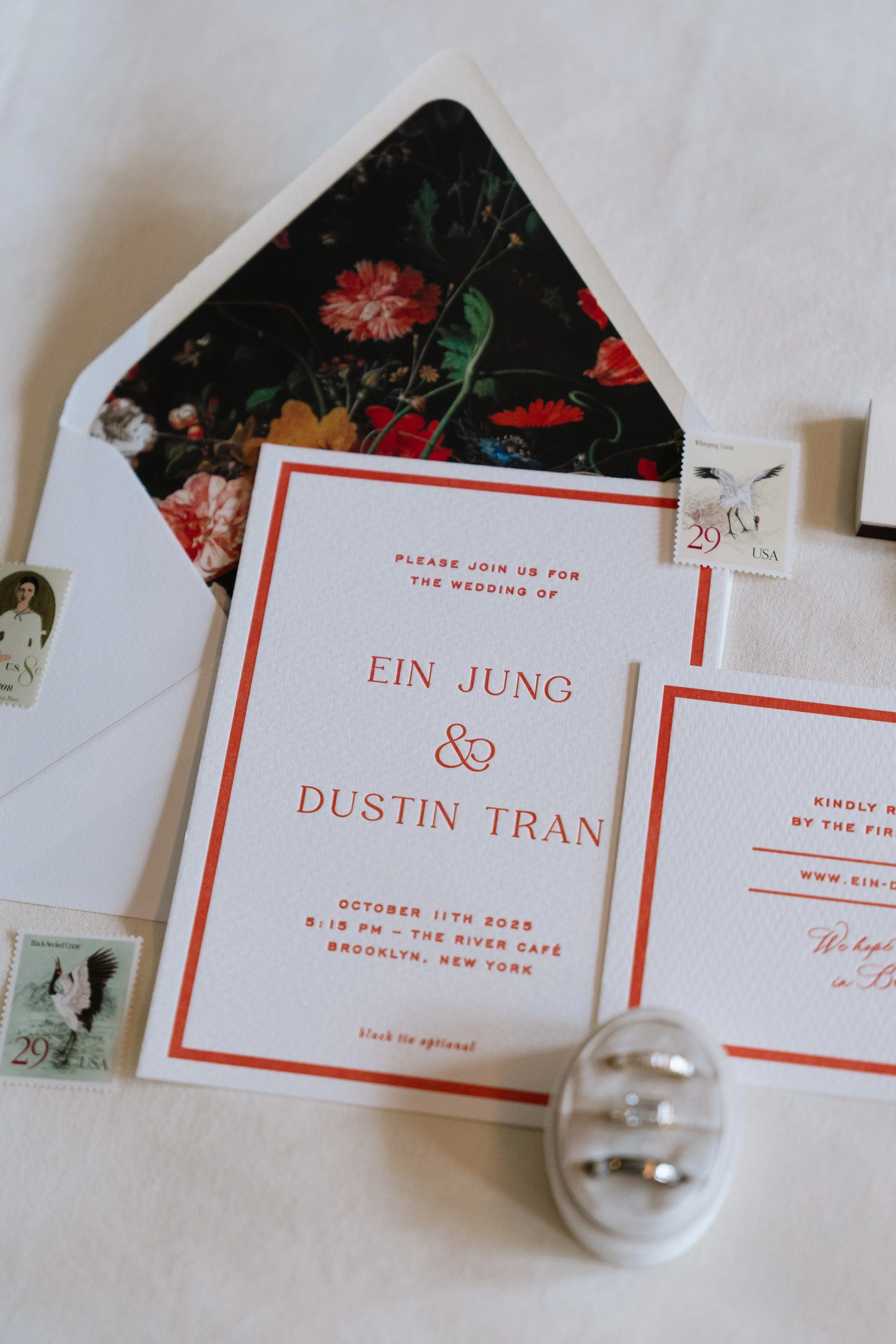 Wedding invitation with details of the wedding of Ein Jung and Dustin Tran, scheduled for October 11th, 2025 at 5:15 PM at The River Café in Brooklyn, New York, surrounded by wedding accessories and stamps.