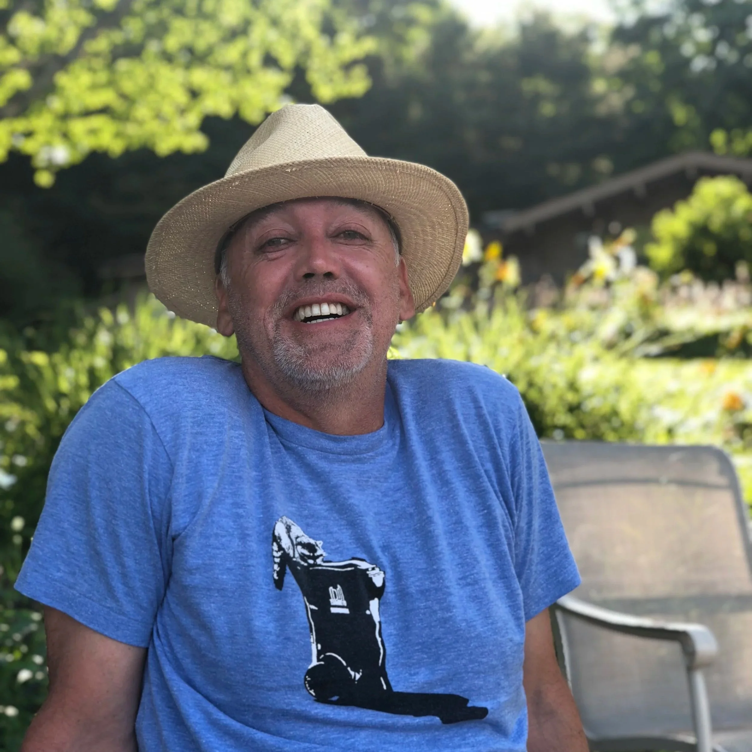 A man wearing a straw hat and a blue t-shirt with a golf bag graphic, sitting outdoors in a garden with greenery in the background, smiling at the camera.