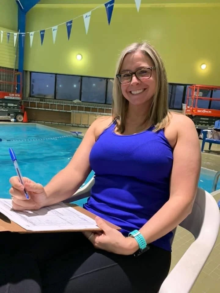 Woman in a blue tank top sitting at a table by a swimming pool, holding a pen and clipboard, smiling, with a watch on her left wrist and glasses, indoors with green walls and blue and white pennant banners overhead.