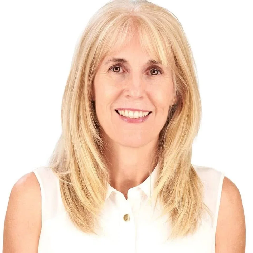 A smiling woman with shoulder-length blonde hair wearing a white sleeveless top against a plain white background.