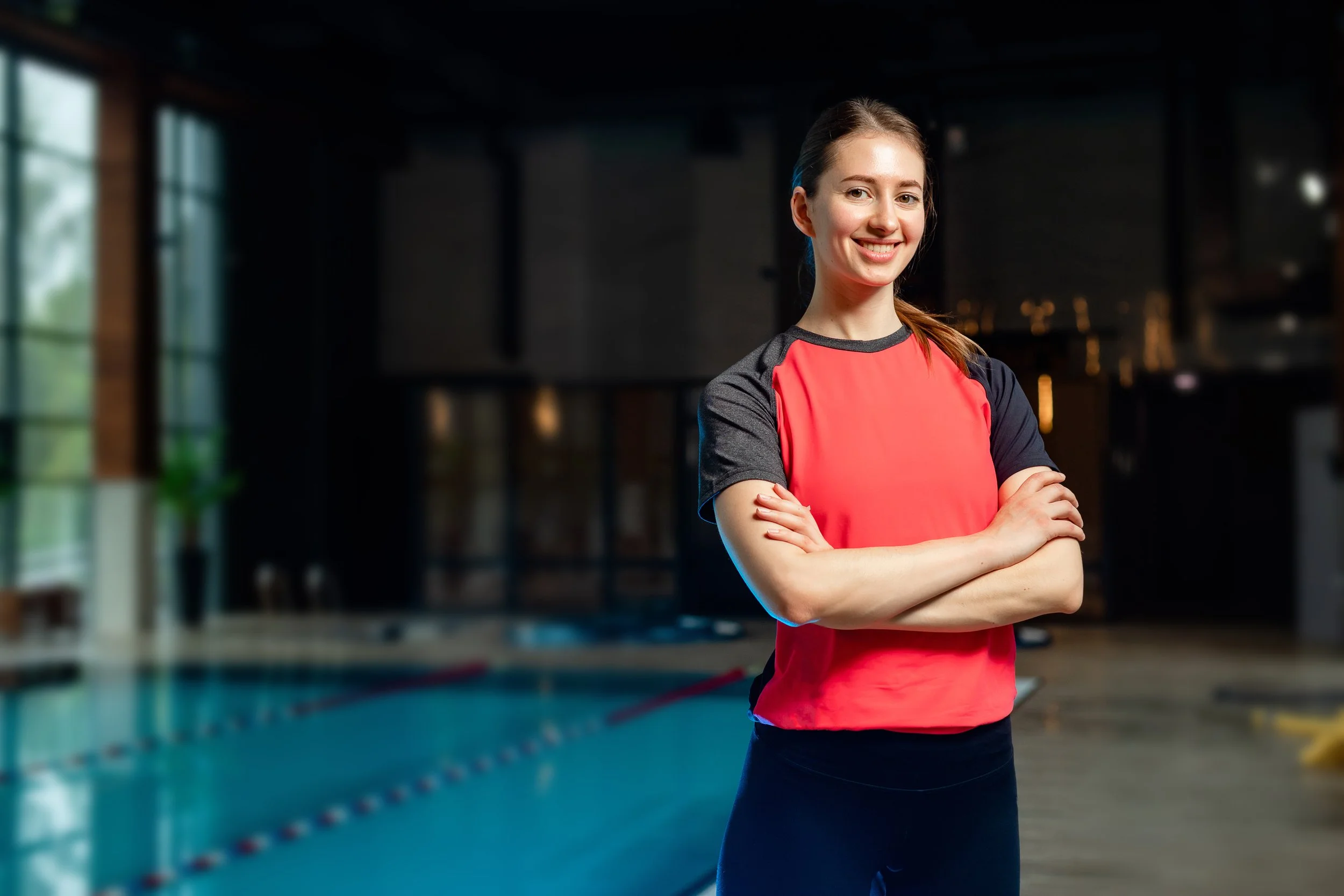 A young woman with long hair tied back, smiling with arms crossed, standing inside an indoor swimming pool facility.