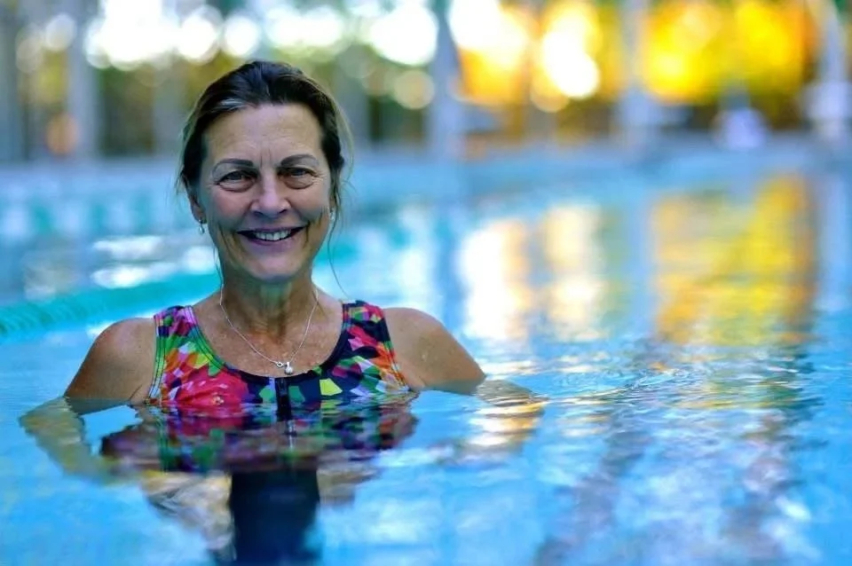 Smiling woman in a colorful swimsuit swimming in an outdoor pool with trees and sunlight in the background.